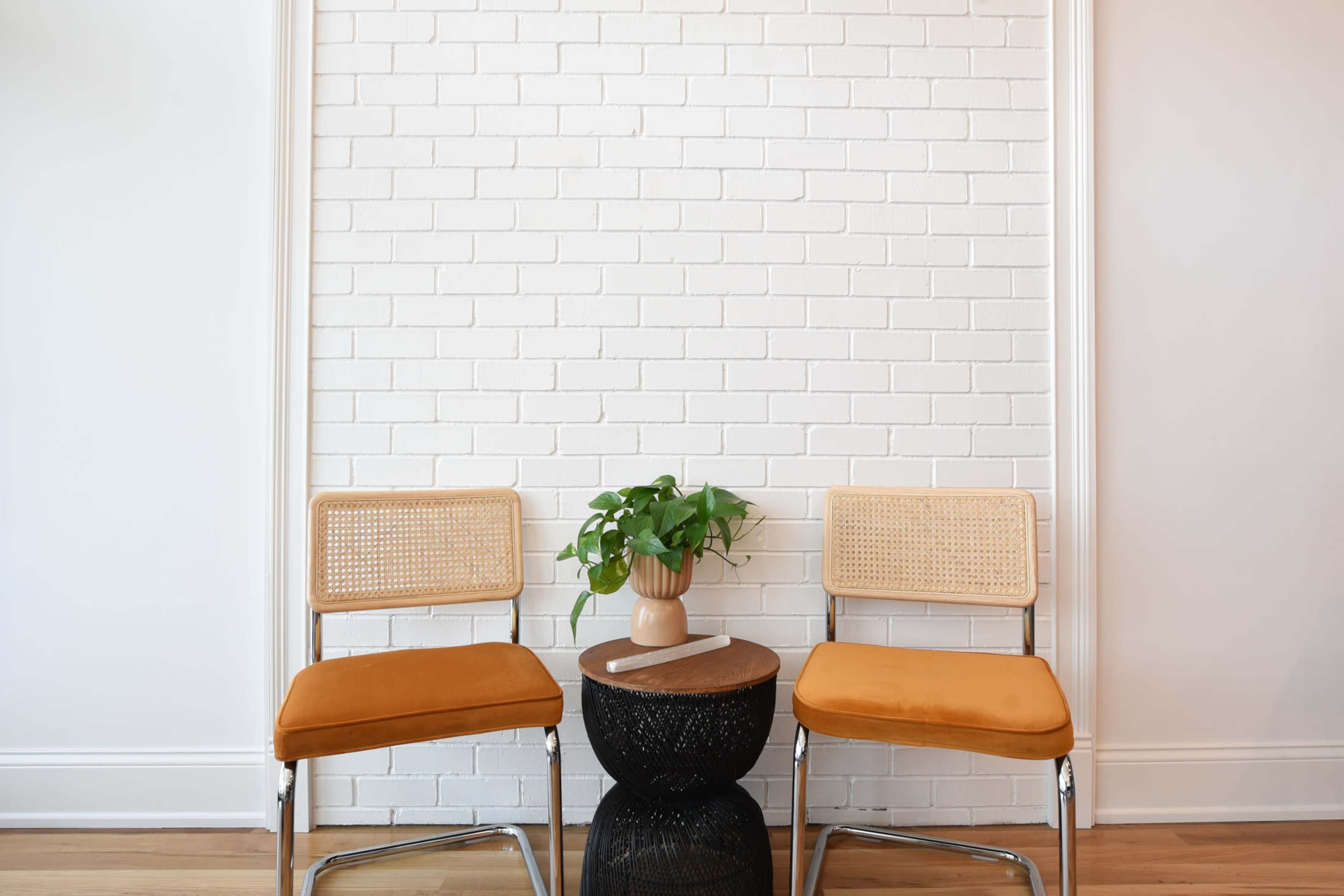 Two orange upholstered chairs are positioned on either side of a small round table with a plant and a ceramic vase, against a white brick wall.