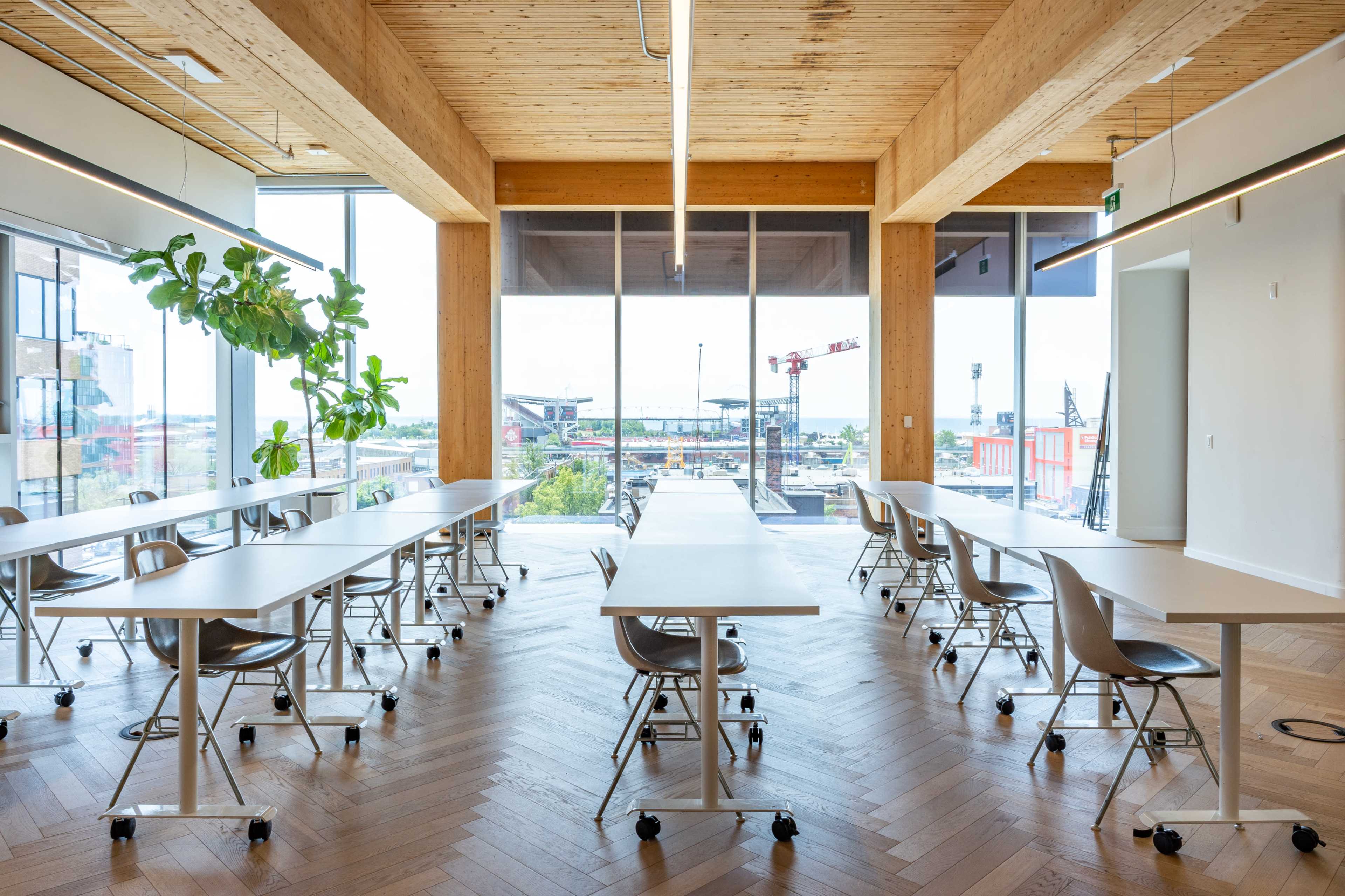 The image shows a modern classroom or meeting space with multiple tables arranged for group work, large windows letting in natural light, and a view of construction equipment outside.