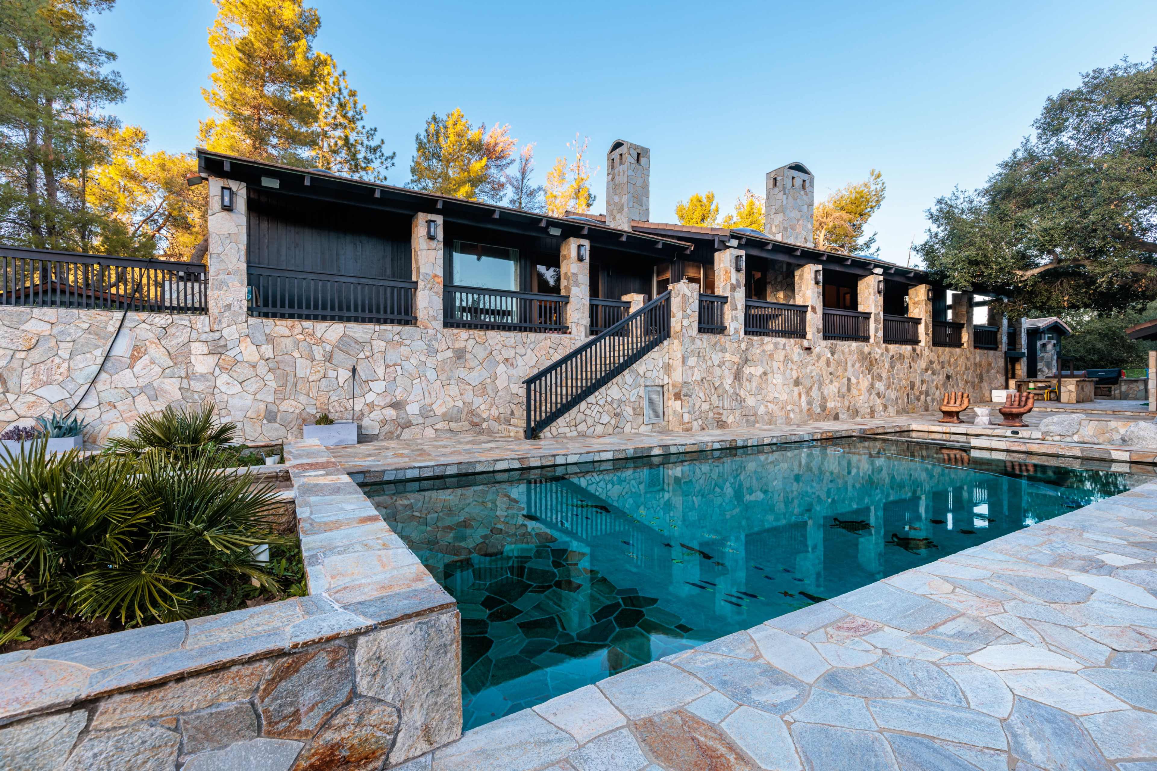 The image shows a stone-lined swimming pool in front of a rustic, two-story house surrounded by trees.