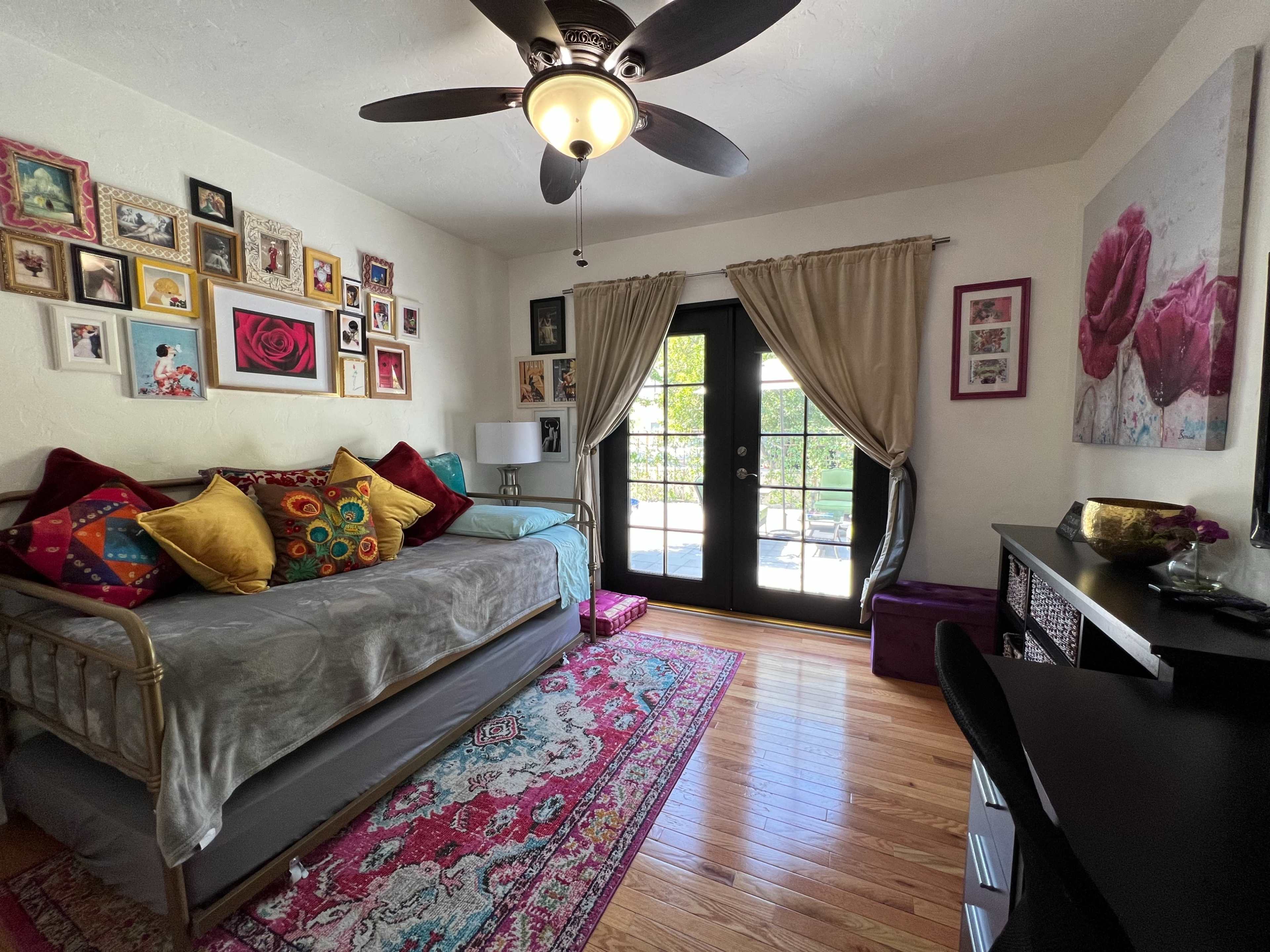 The room features a daybed adorned with colorful pillows, a desk against the wall, and large windows with double doors leading to an outdoor space.