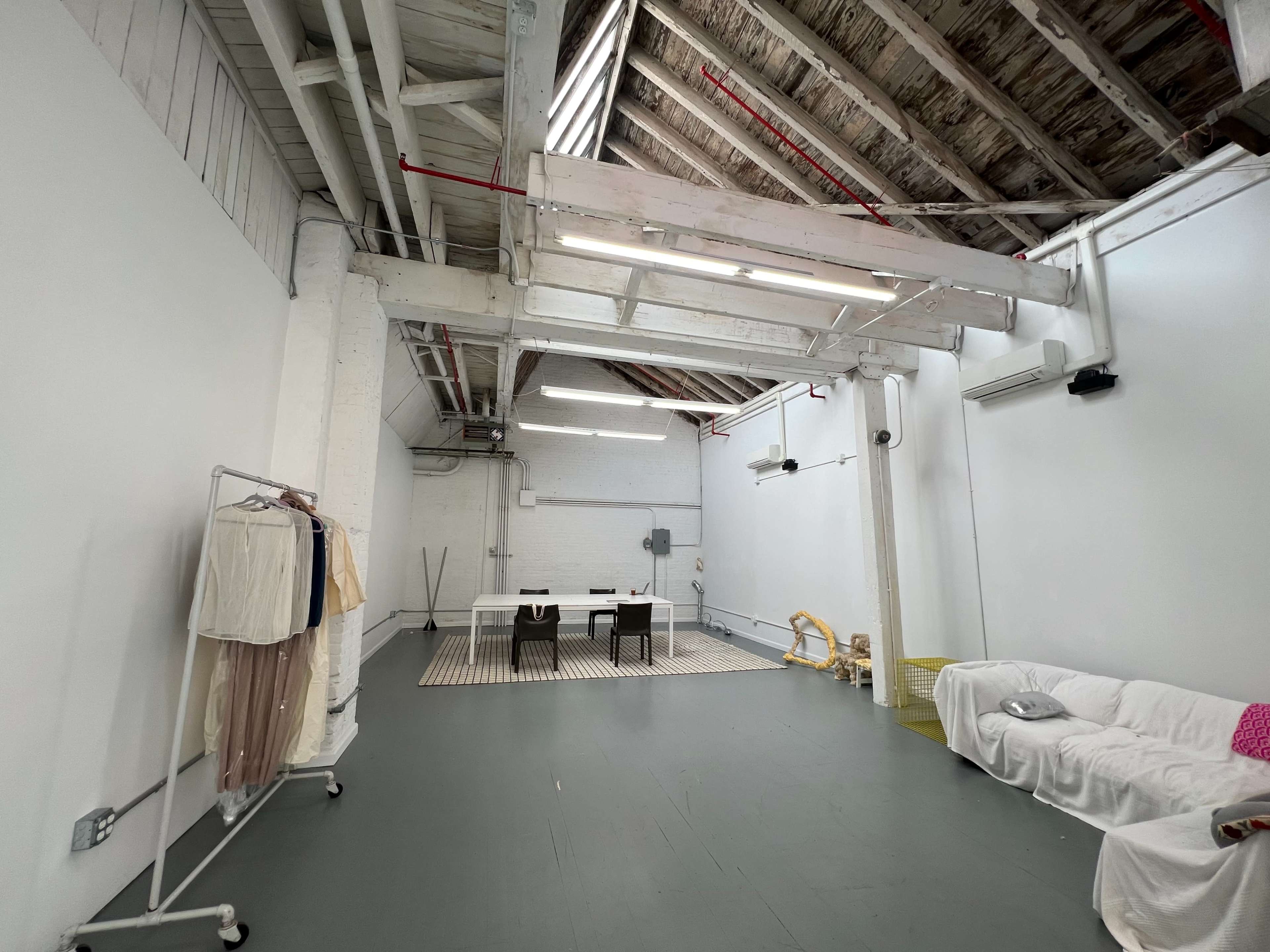 A spacious, minimalist interior with exposed beams, a large table, chairs, and a clothing rack against the white walls.