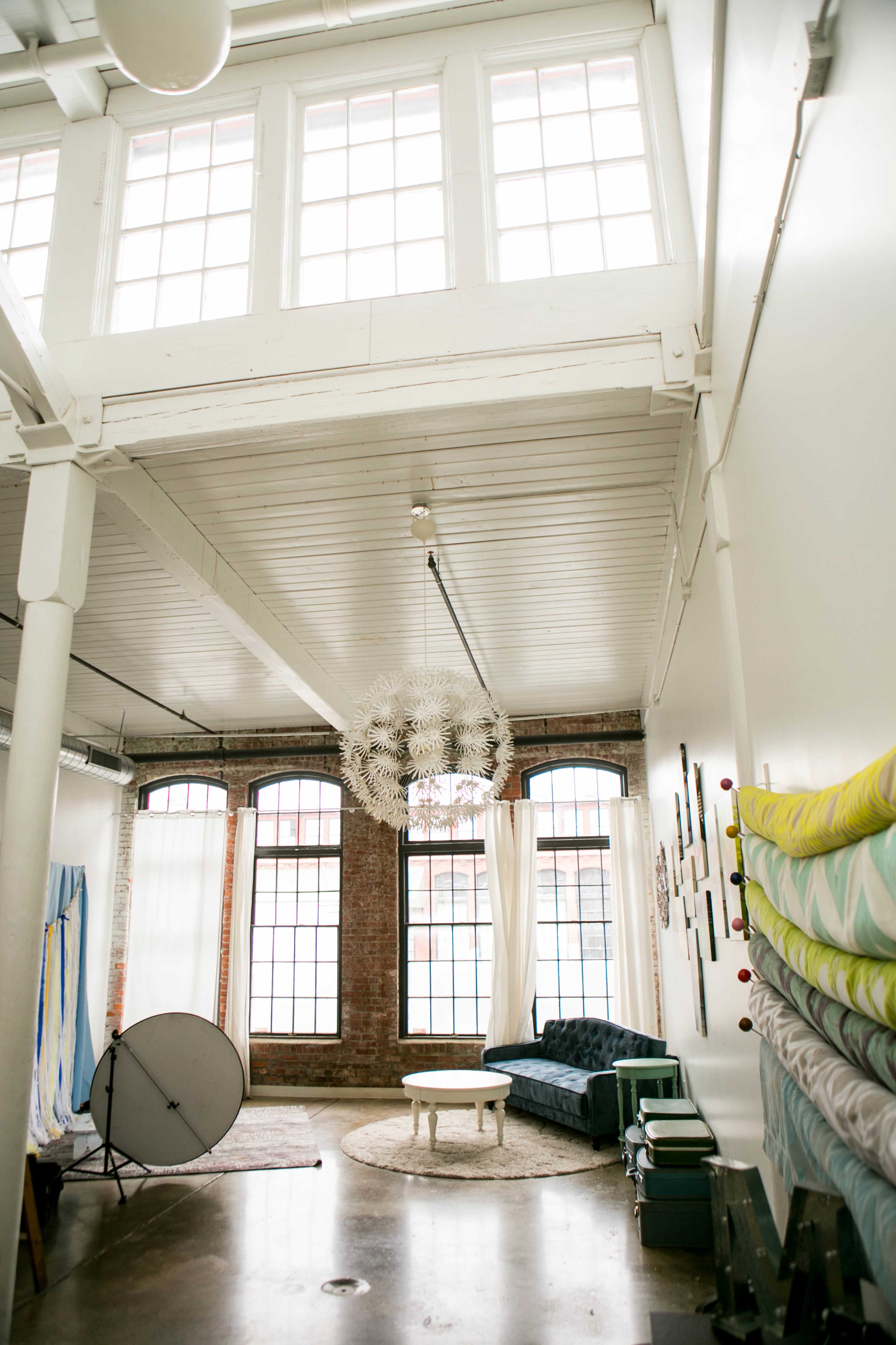 A spacious, well-lit interior with a high ceiling, large windows, and a central circular rug beneath a coffee table, surrounded by a sectional sofa and decorative wall hangings.