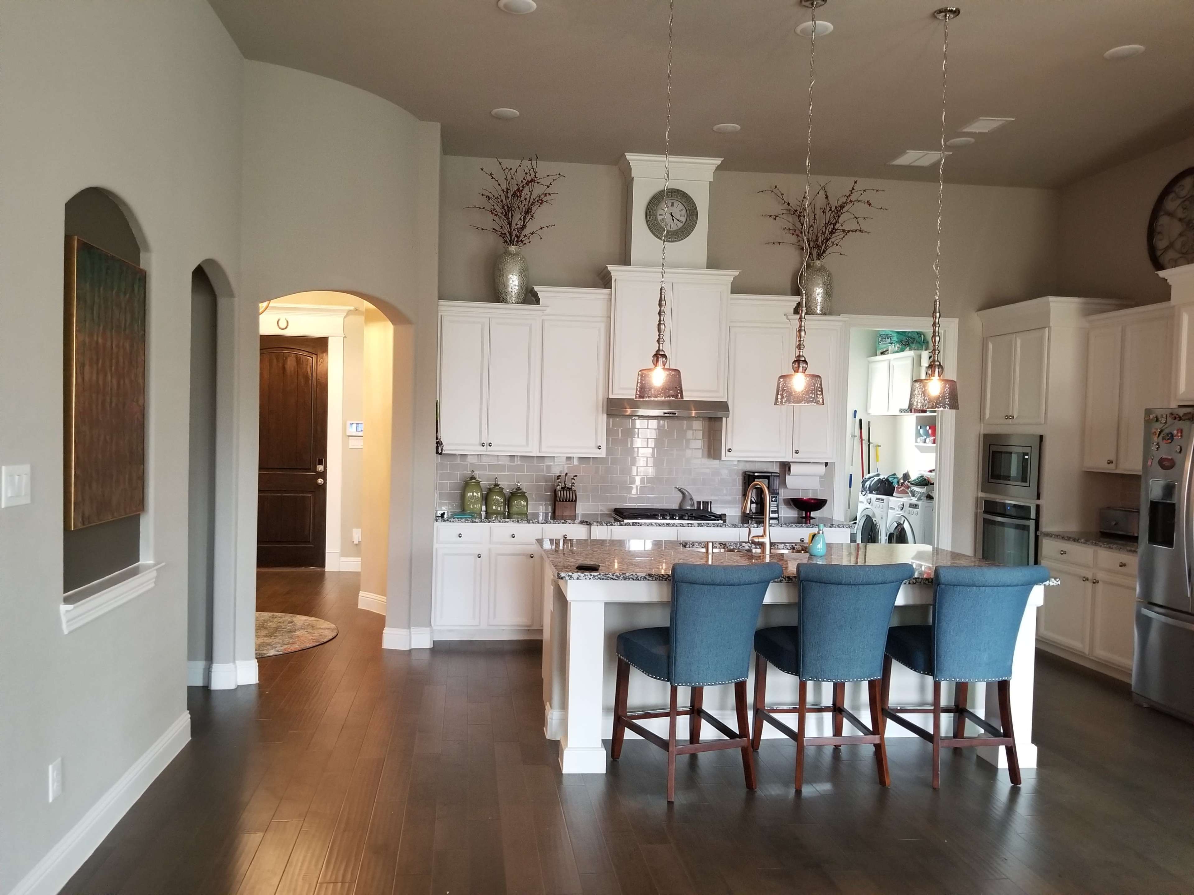 A modern kitchen features white cabinetry, a central island with blue bar stools, and stainless steel appliances, with an adjacent dining area and entryway.