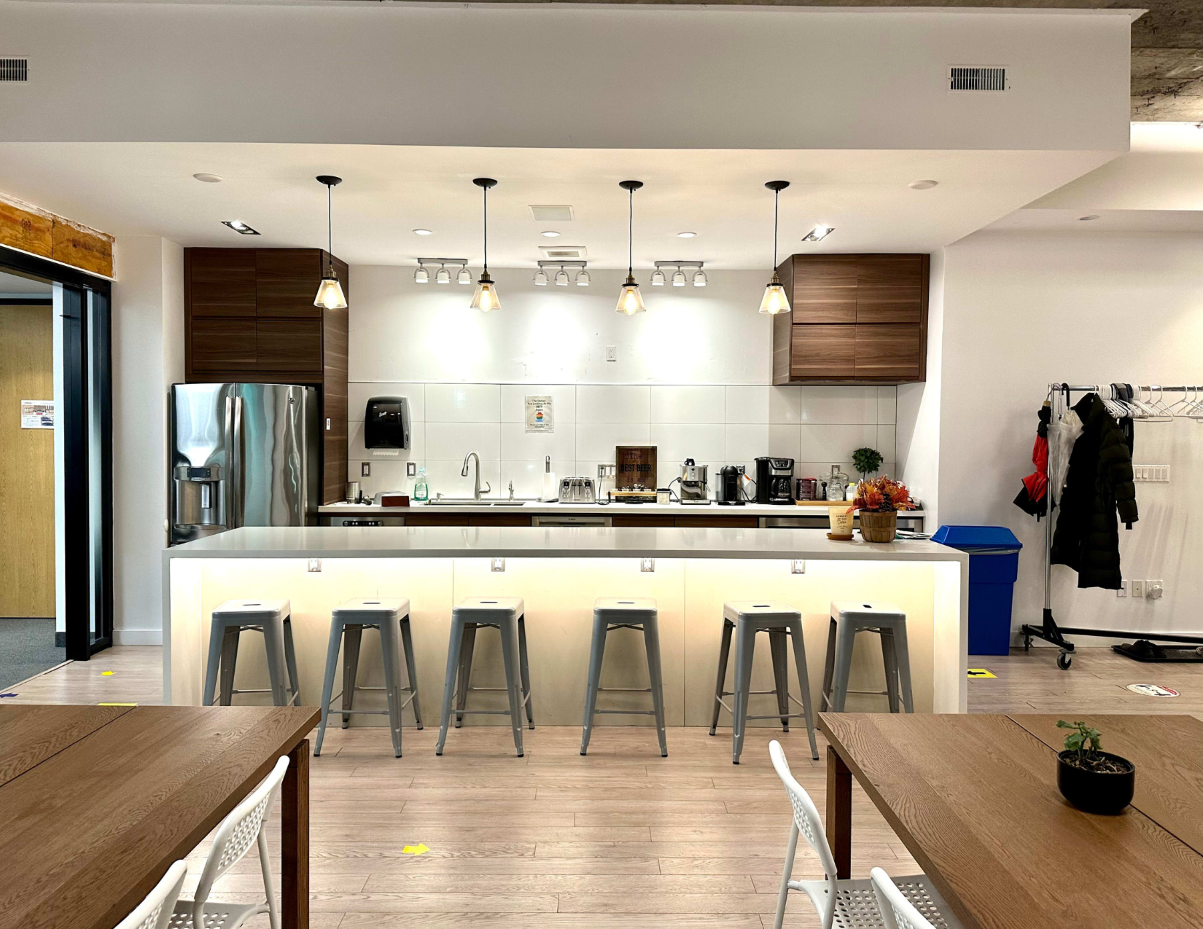 The image shows a modern kitchen area with a bar counter, stools, and various appliances, including a refrigerator and coffee machines.