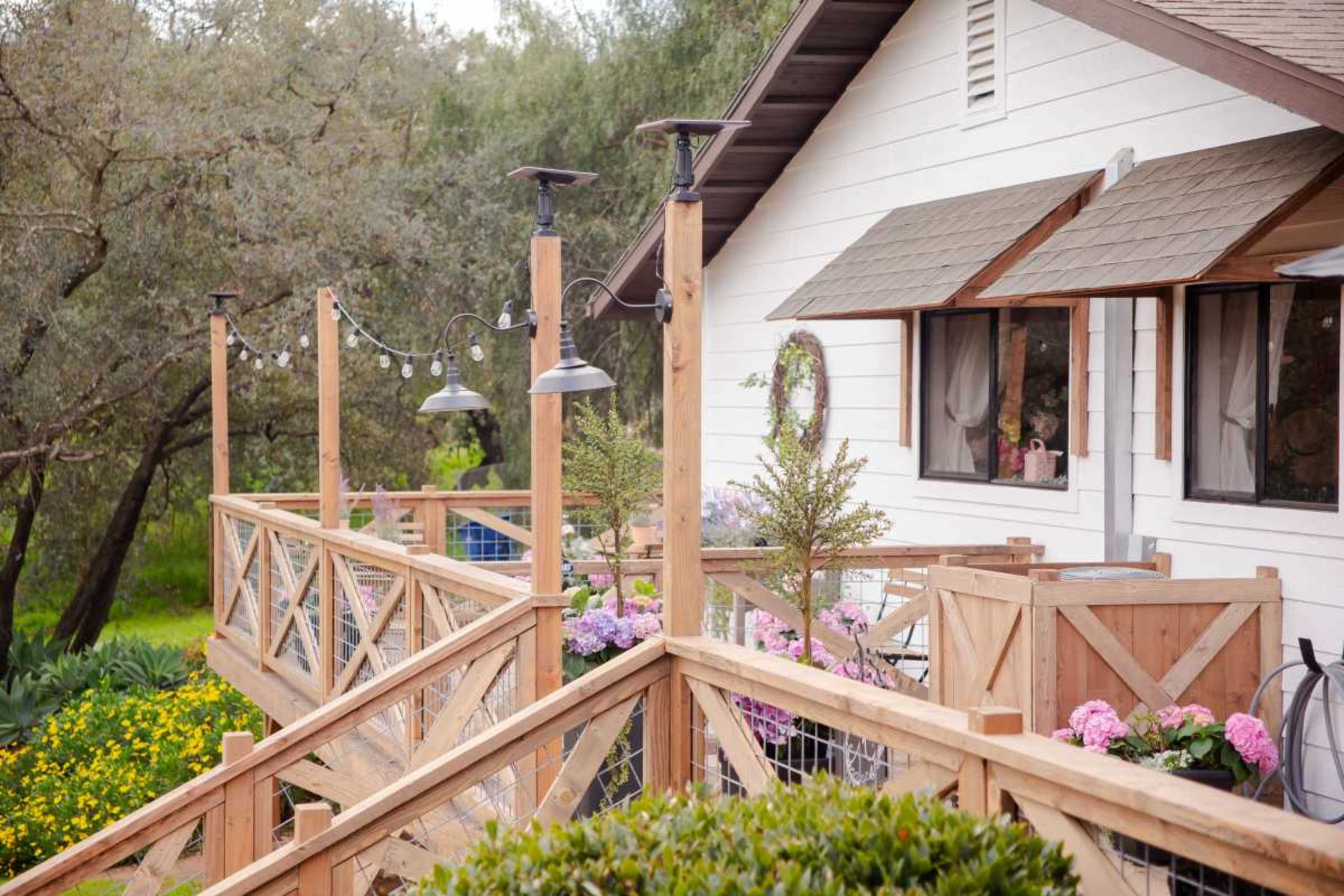 A wooden deck surrounded by flowering plants, leading to a house with a light-colored exterior and multiple windows.
