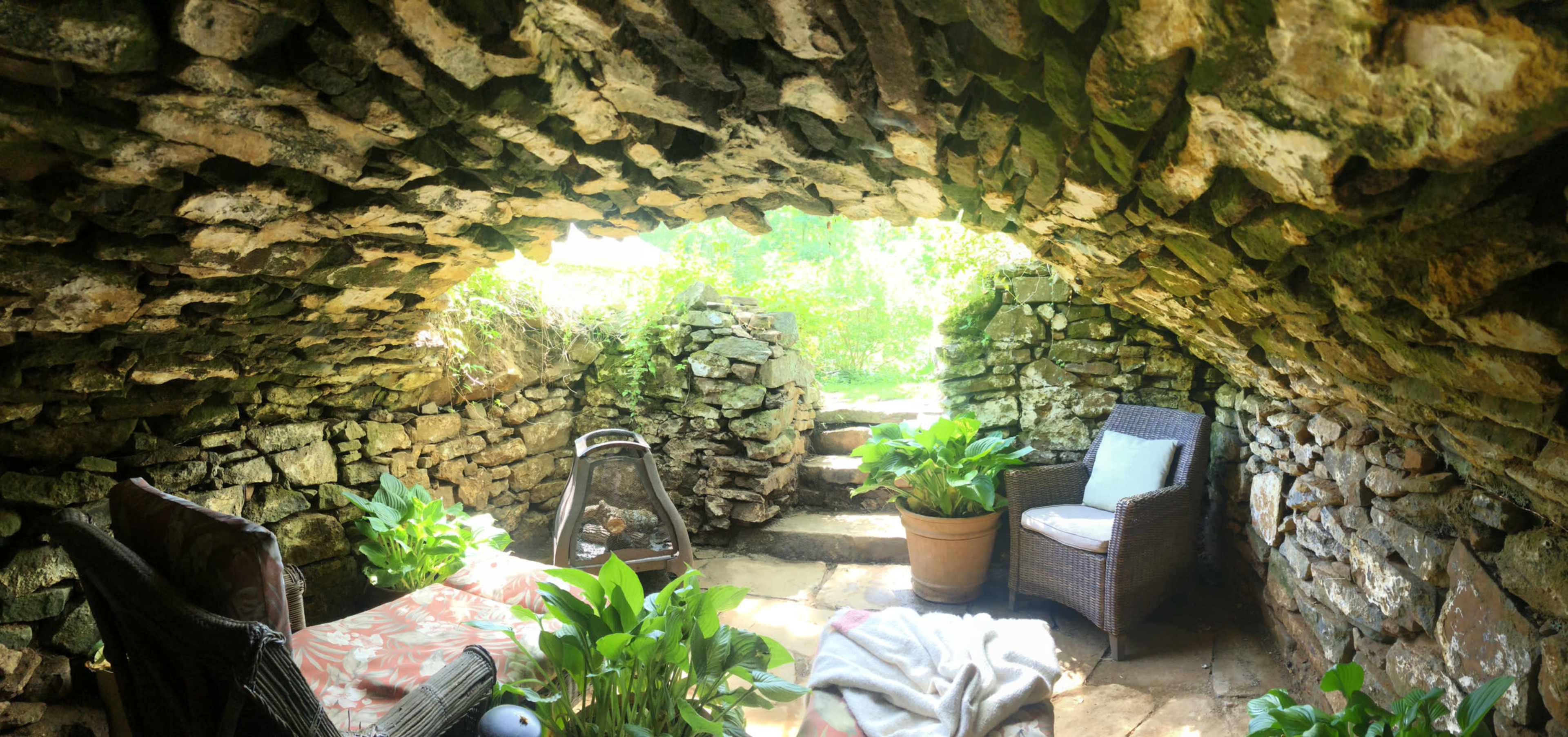 The image shows a stone-walled alcove with two chairs, potted plants, and natural light filtering in through the open entrance.