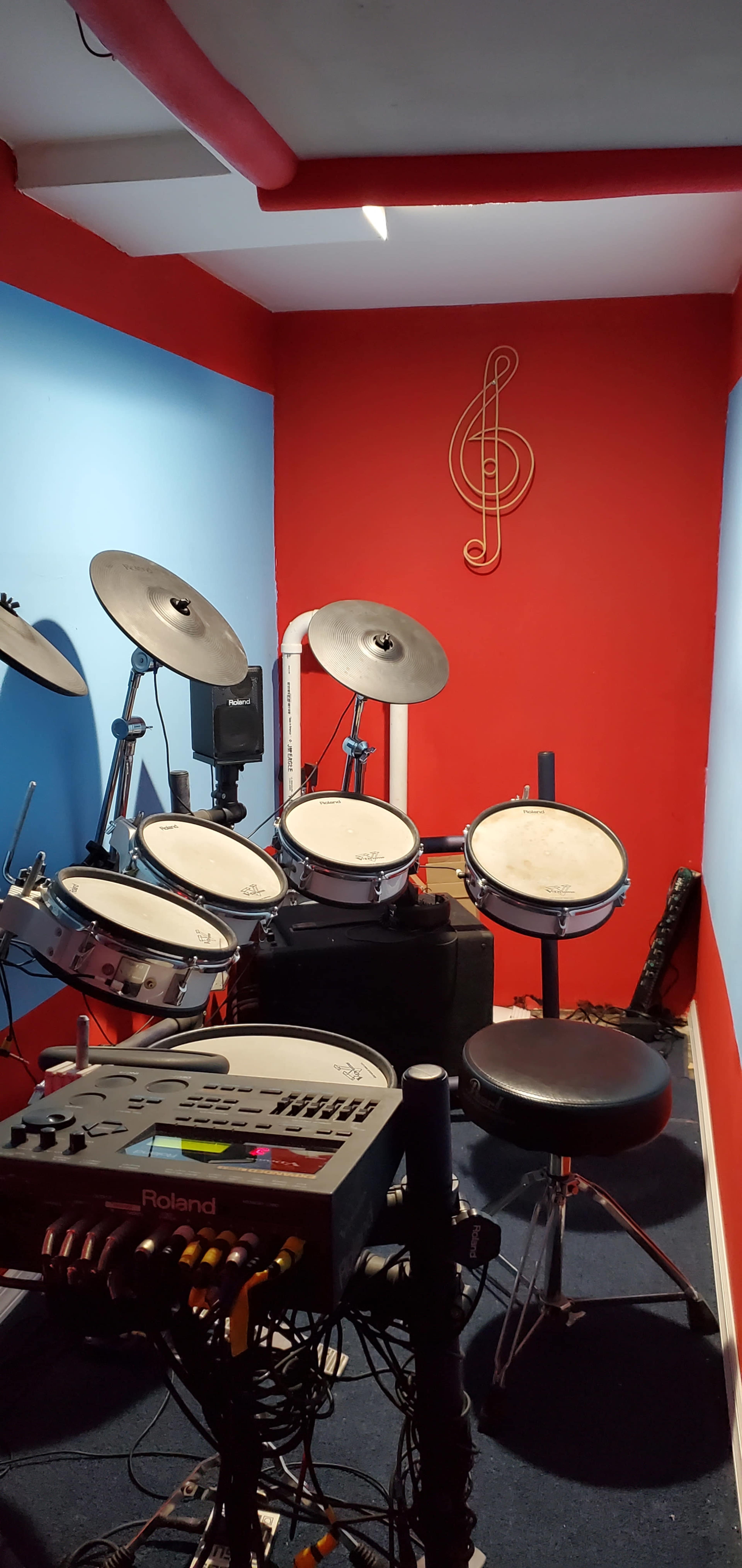 A small music practice room features an electronic drum set against a red and blue wall, with a musical clef decoration.