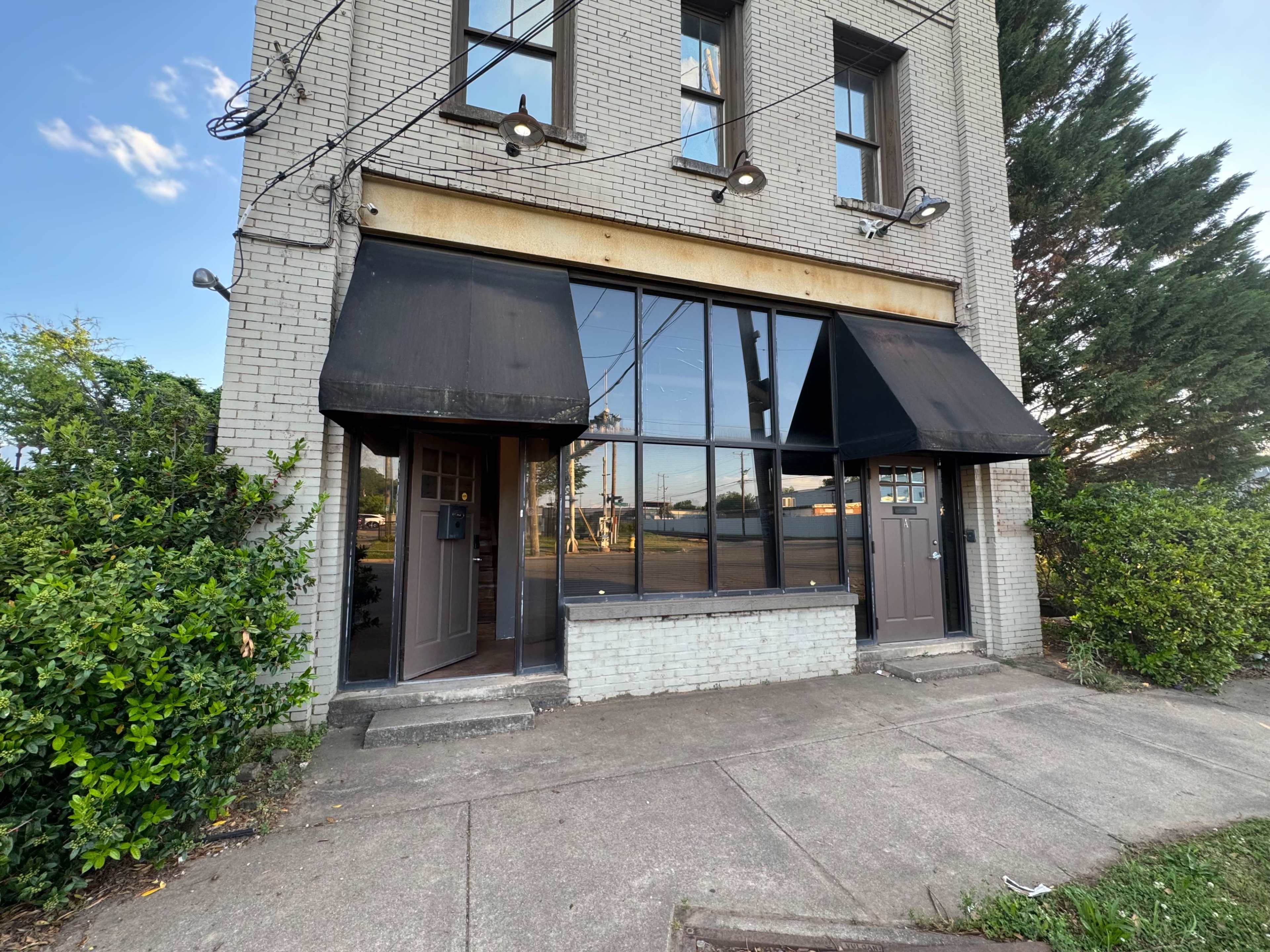 The image shows a gray brick building with two doors and large windows, each covered by black awnings, situated on a sidewalk surrounded by greenery.
