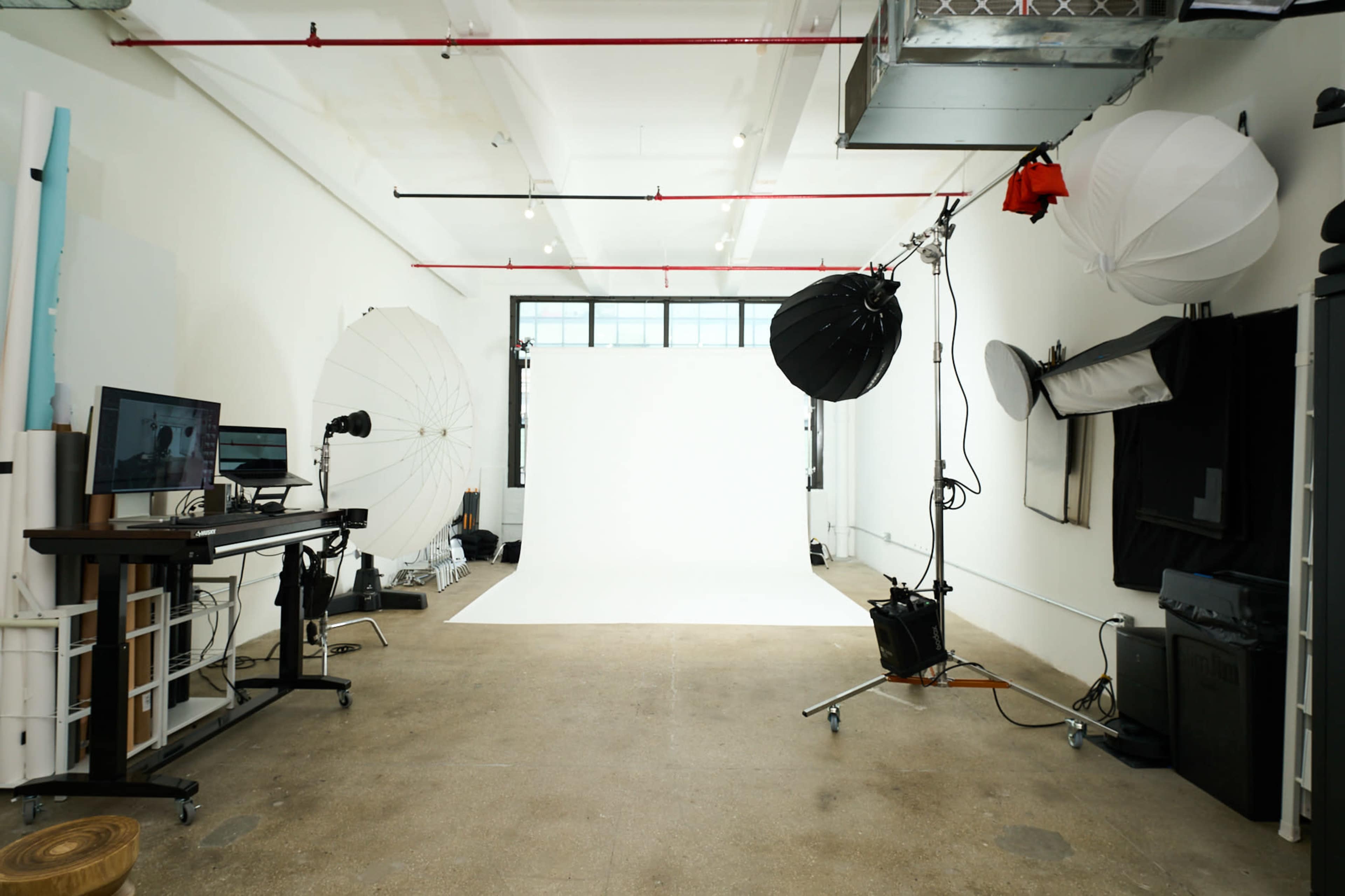 A photography studio features a white backdrop, various lighting equipment, and a workspace with a monitor.