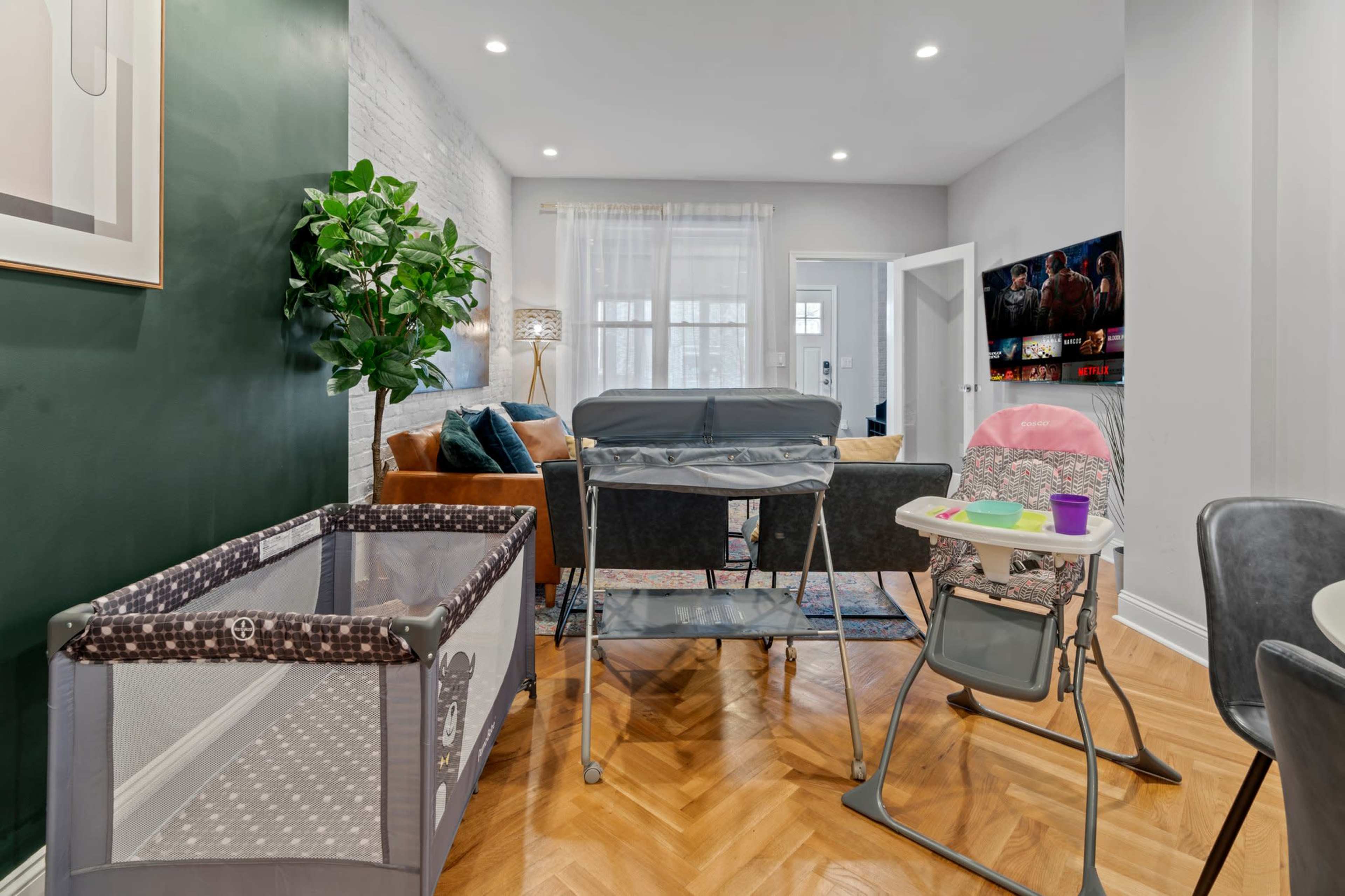 A bright living room featuring a playpen, a bassinet, and a high chair, with a couch and TV in the background.