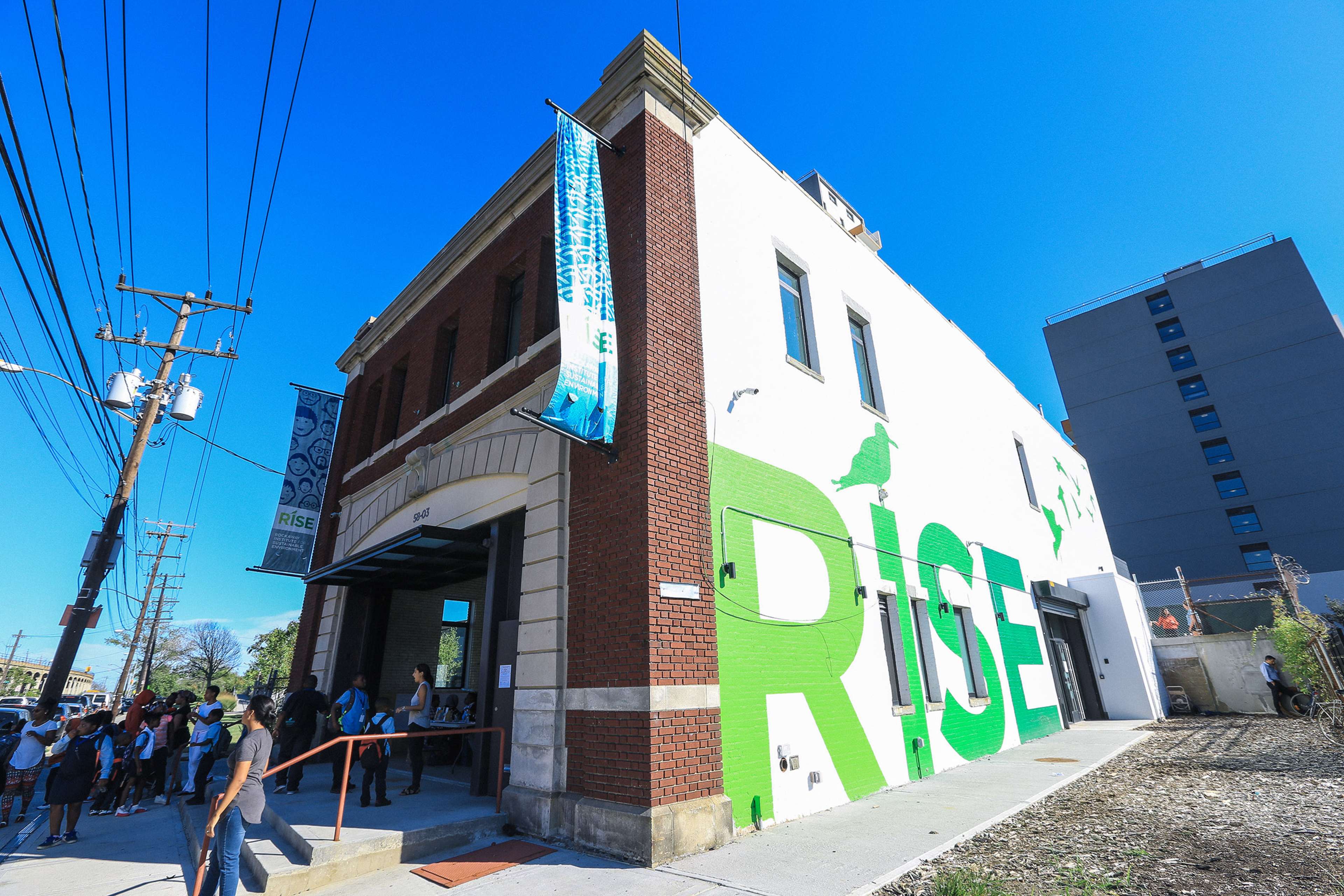 A brick building with a large "RISE" mural on its side, surrounded by a line of people waiting outside.