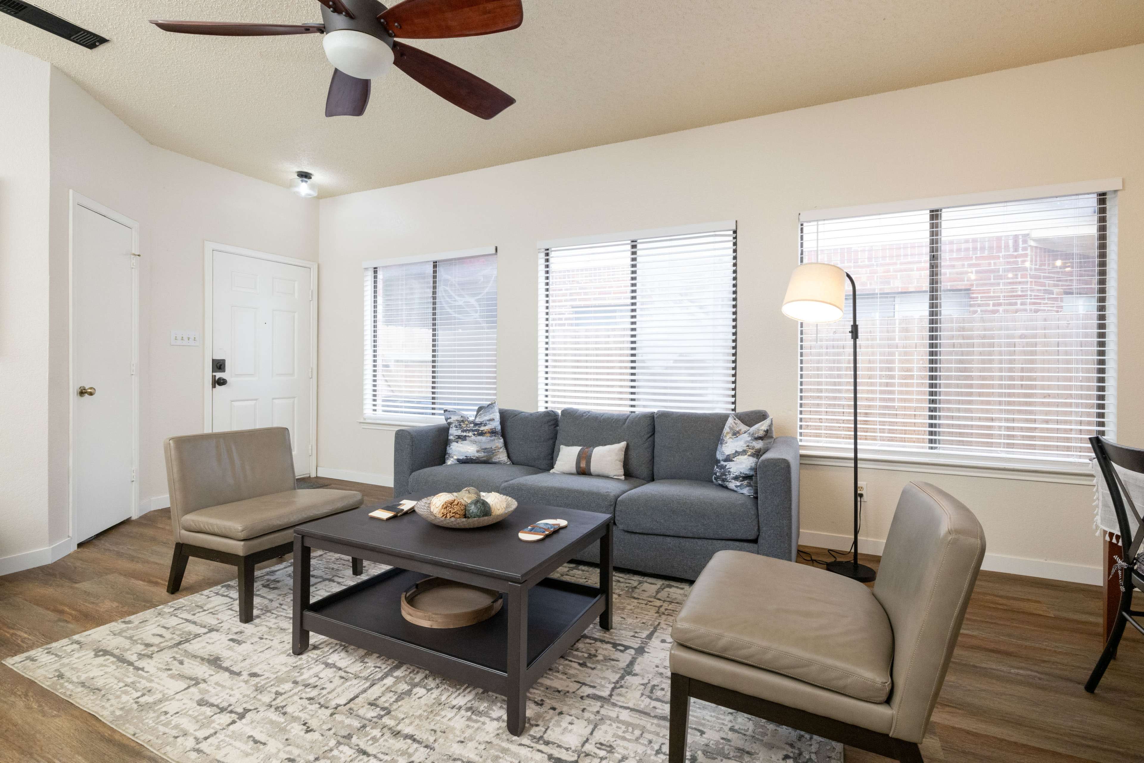 The image shows a modern living room featuring a gray sofa, two matching chairs, a dark coffee table, and large windows with blinds.