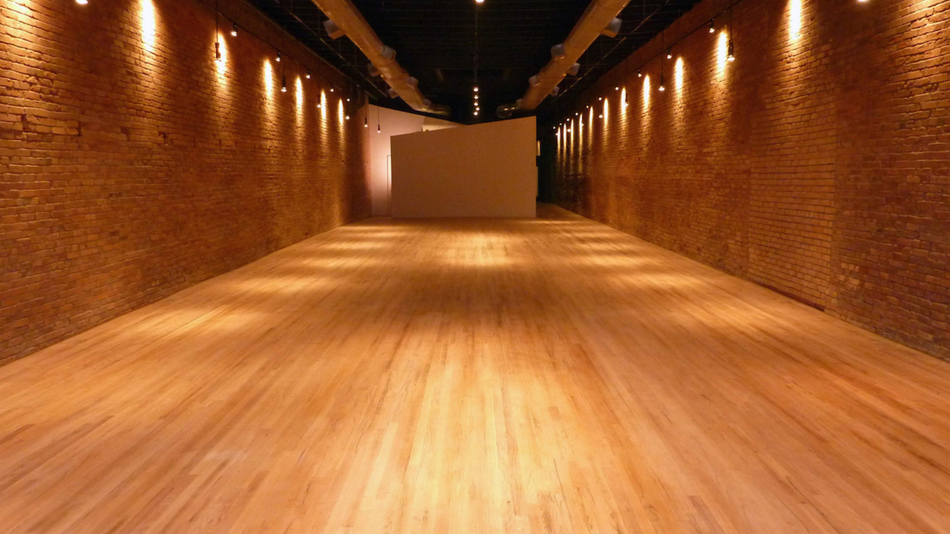 A long, narrow gallery space with exposed brick walls and wooden flooring, illuminated by soft overhead lights.