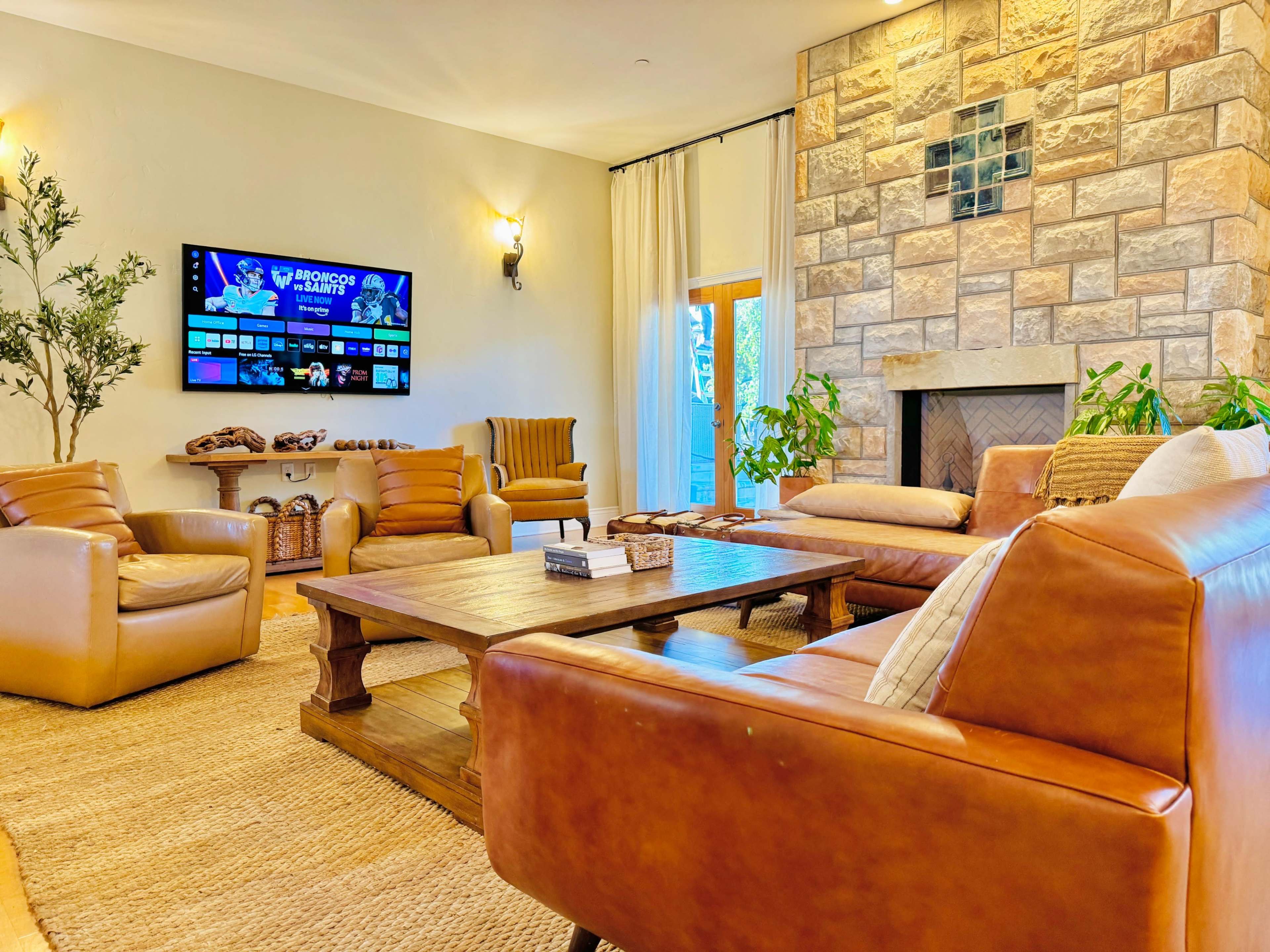 The image depicts a cozy living room with leather sofas, armchairs, a wooden coffee table, a stone fireplace, and a television mounted on the wall.
