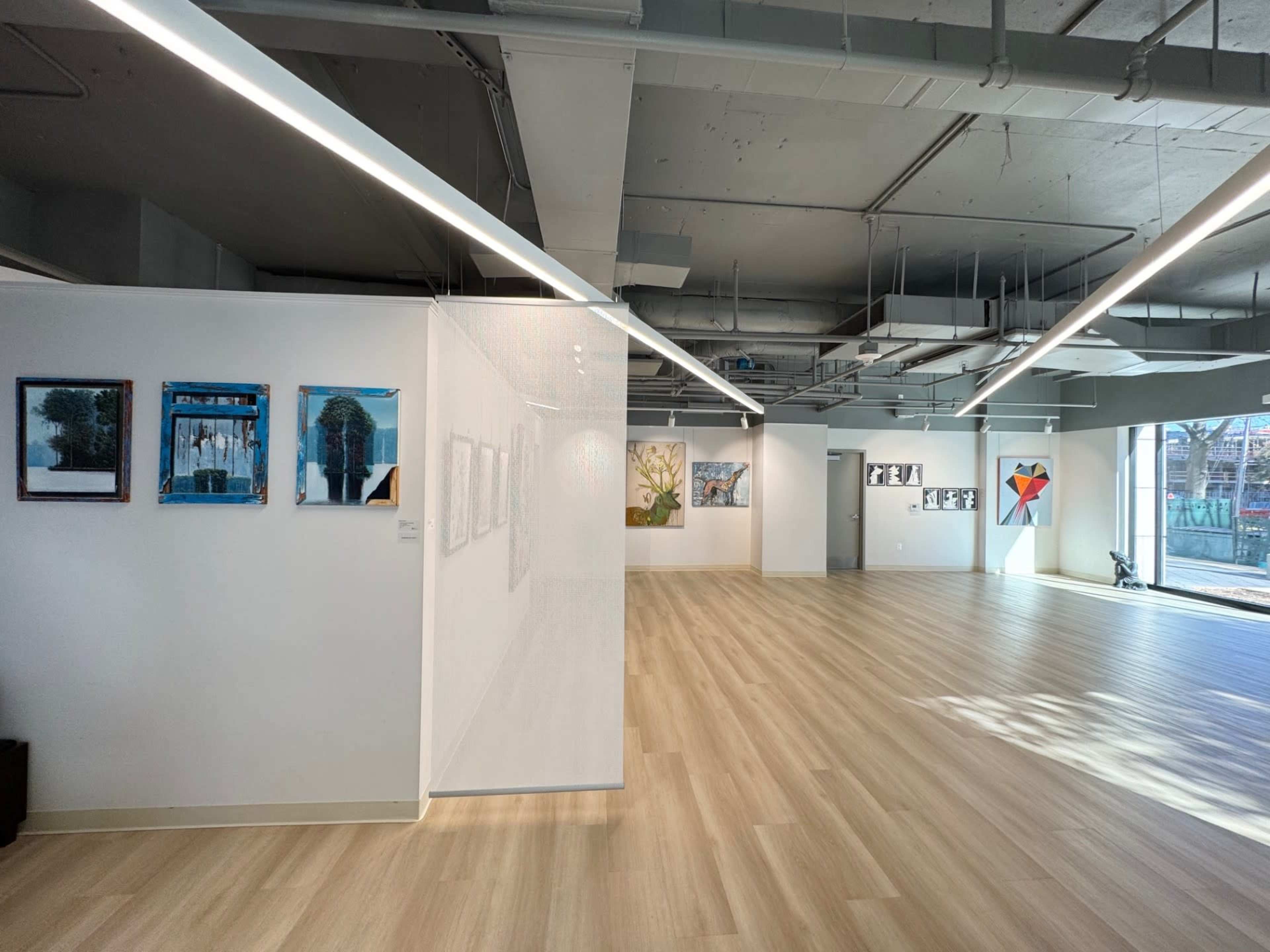 The image shows a spacious art gallery with various framed artworks displayed on the walls and a light-colored wooden floor.