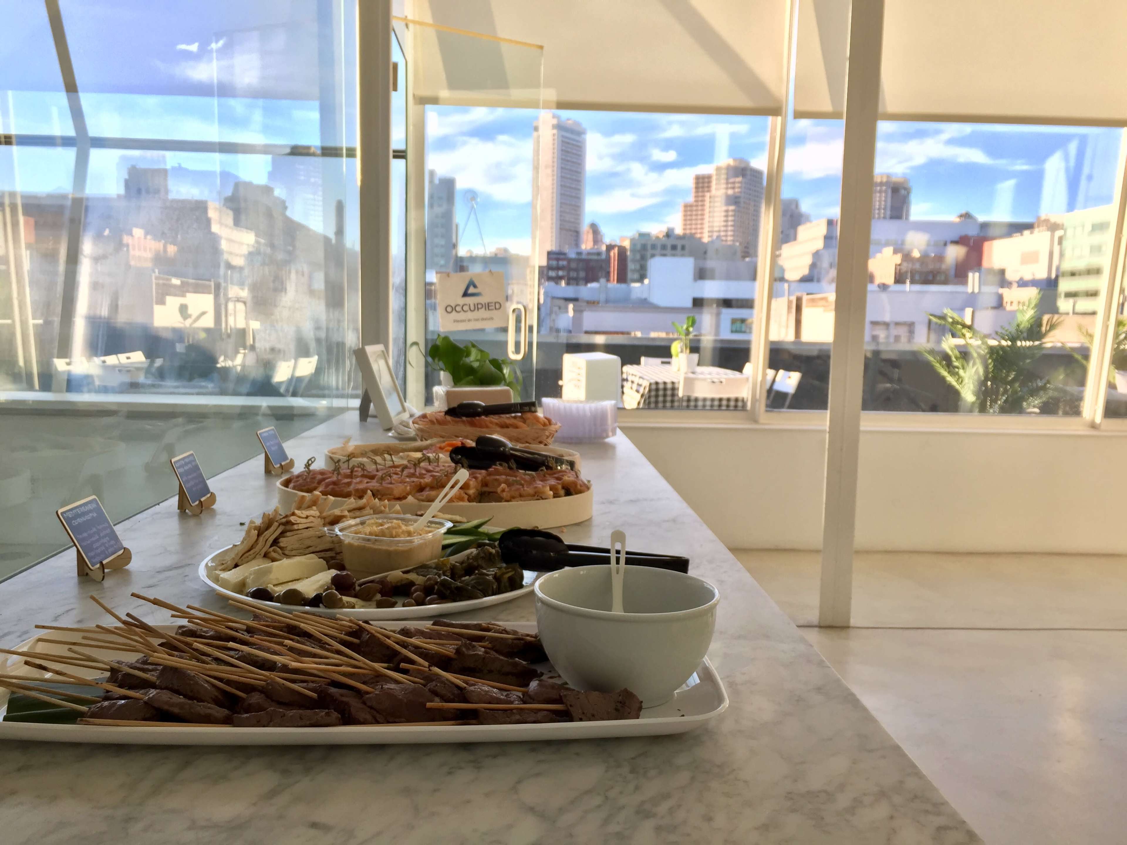 A long marble table is set with various platters of food, including skewers and seafood, overlooking a city skyline through large glass windows.