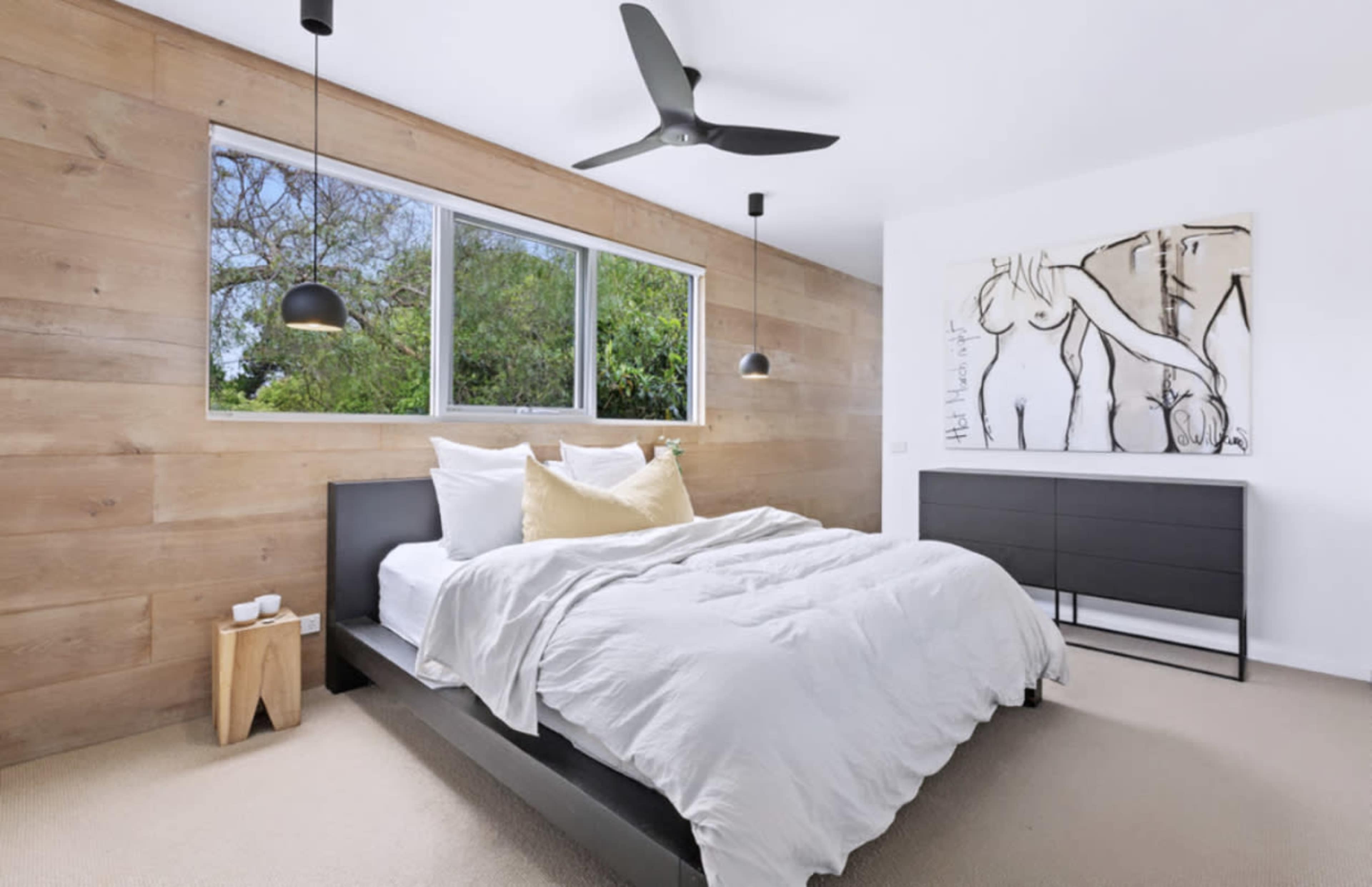 A modern bedroom with a bed covered in white bedding, a ceiling fan, and a large window overlooking greenery.
