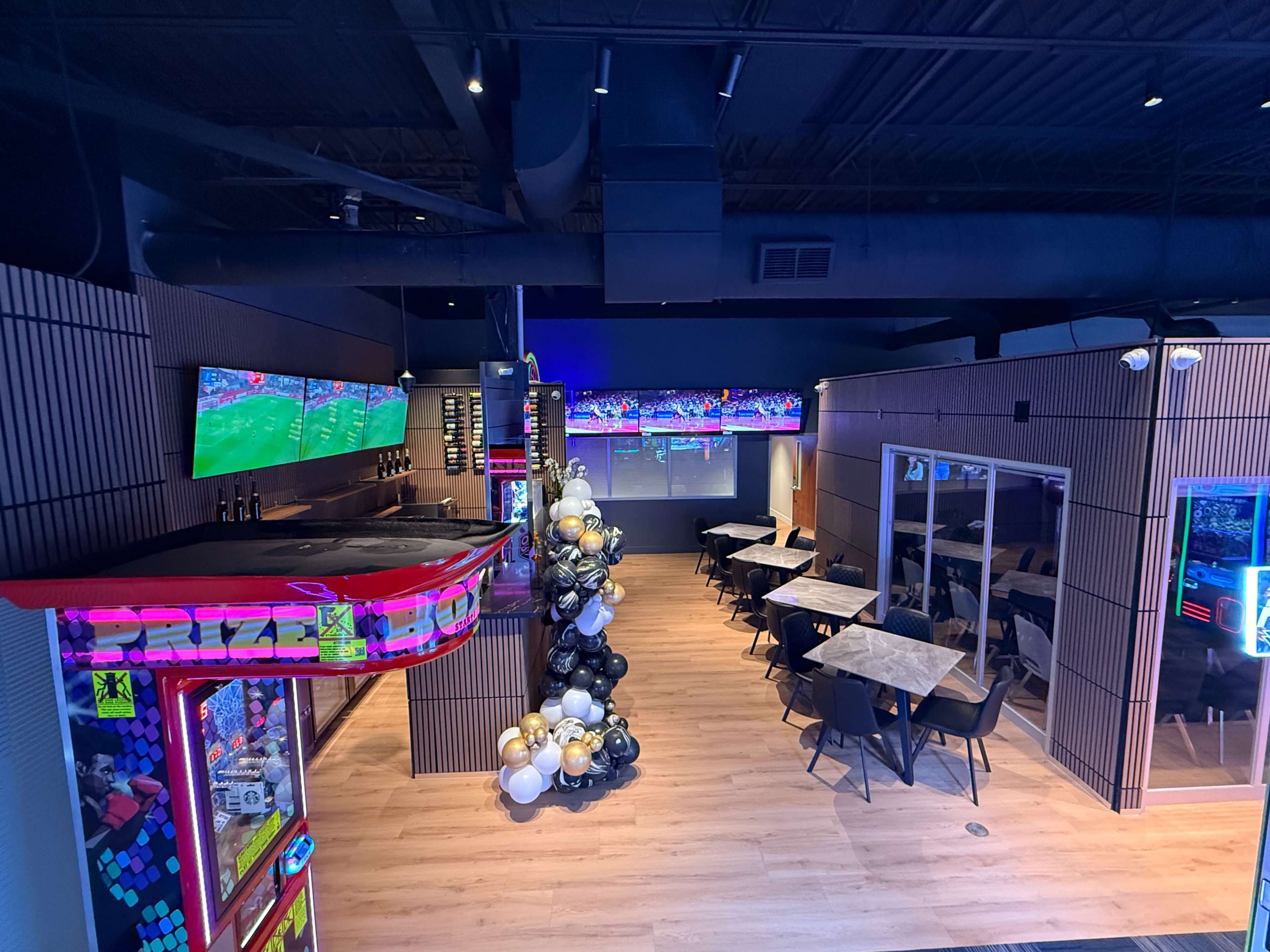 The image shows a modern entertainment space featuring gaming machines, multiple television screens displaying sports, and a dining area with tables and chairs.