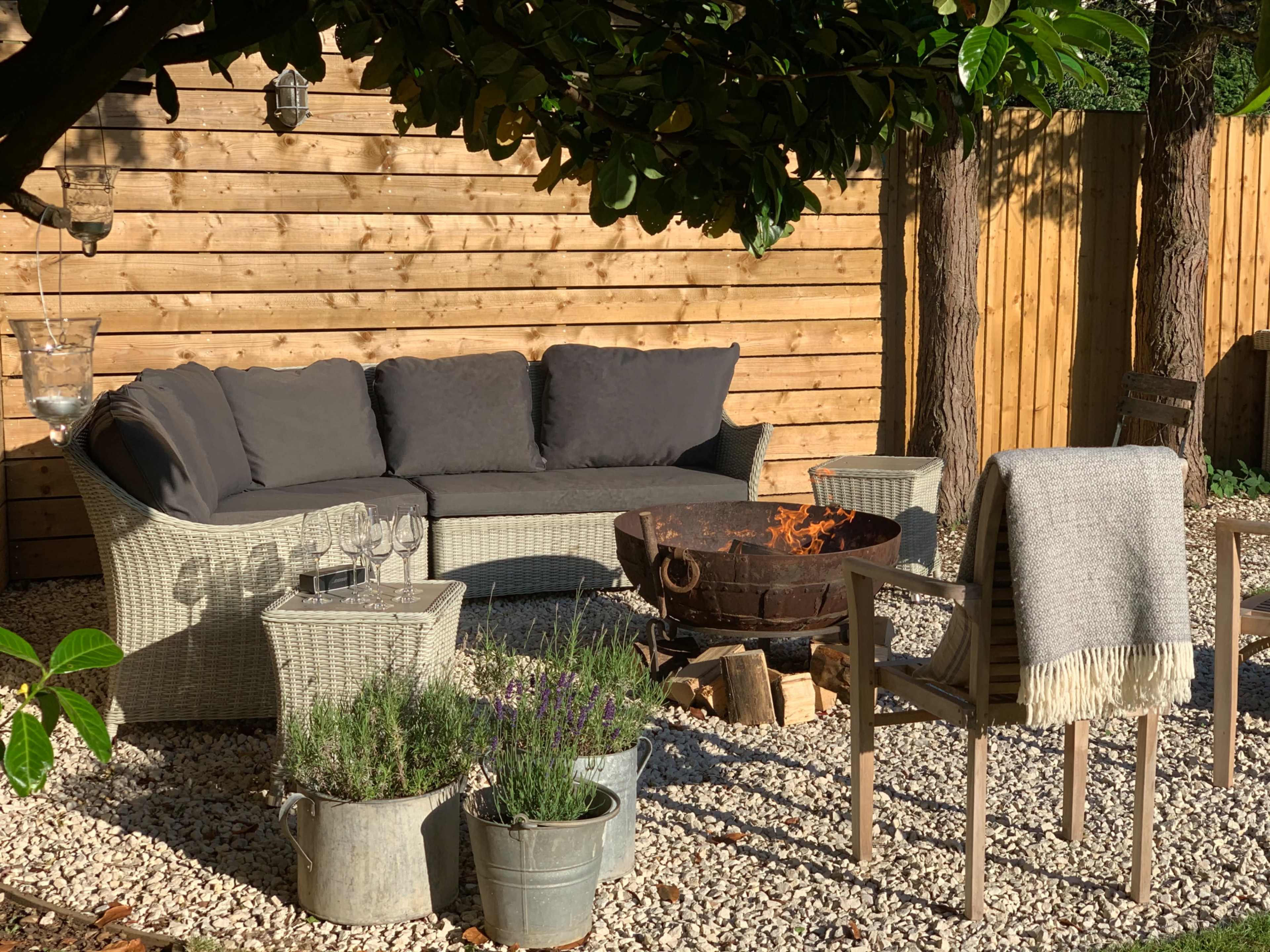 A seating area with a gray sofa, a fire pit, and nearby plants sits on gravel beneath a wooden fence and trees.