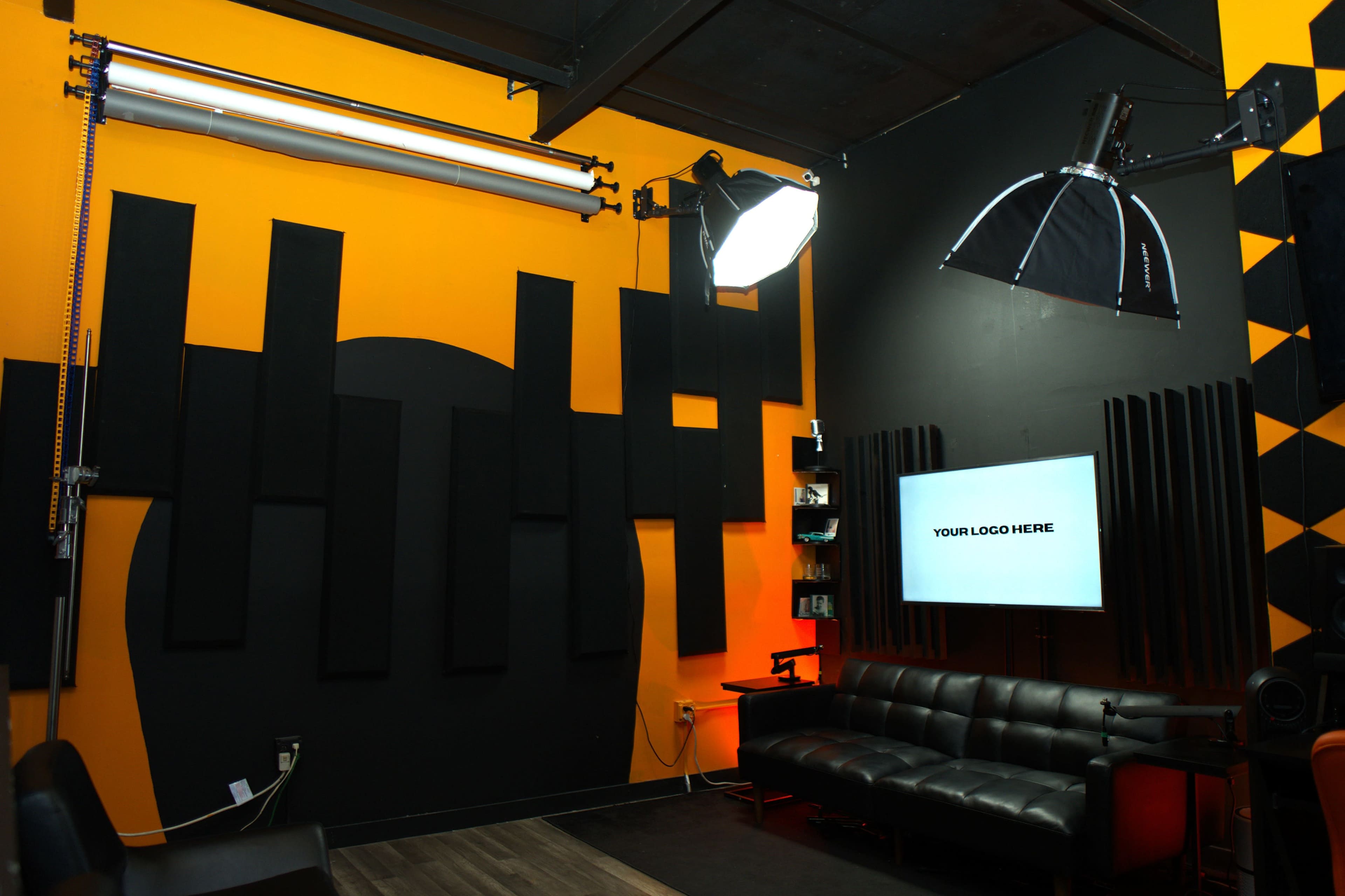 The image shows a modern studio space with orange and black walls, featuring black sound panels, a sleek black sofa, and a television displaying the text "YOUR LOGO HERE."