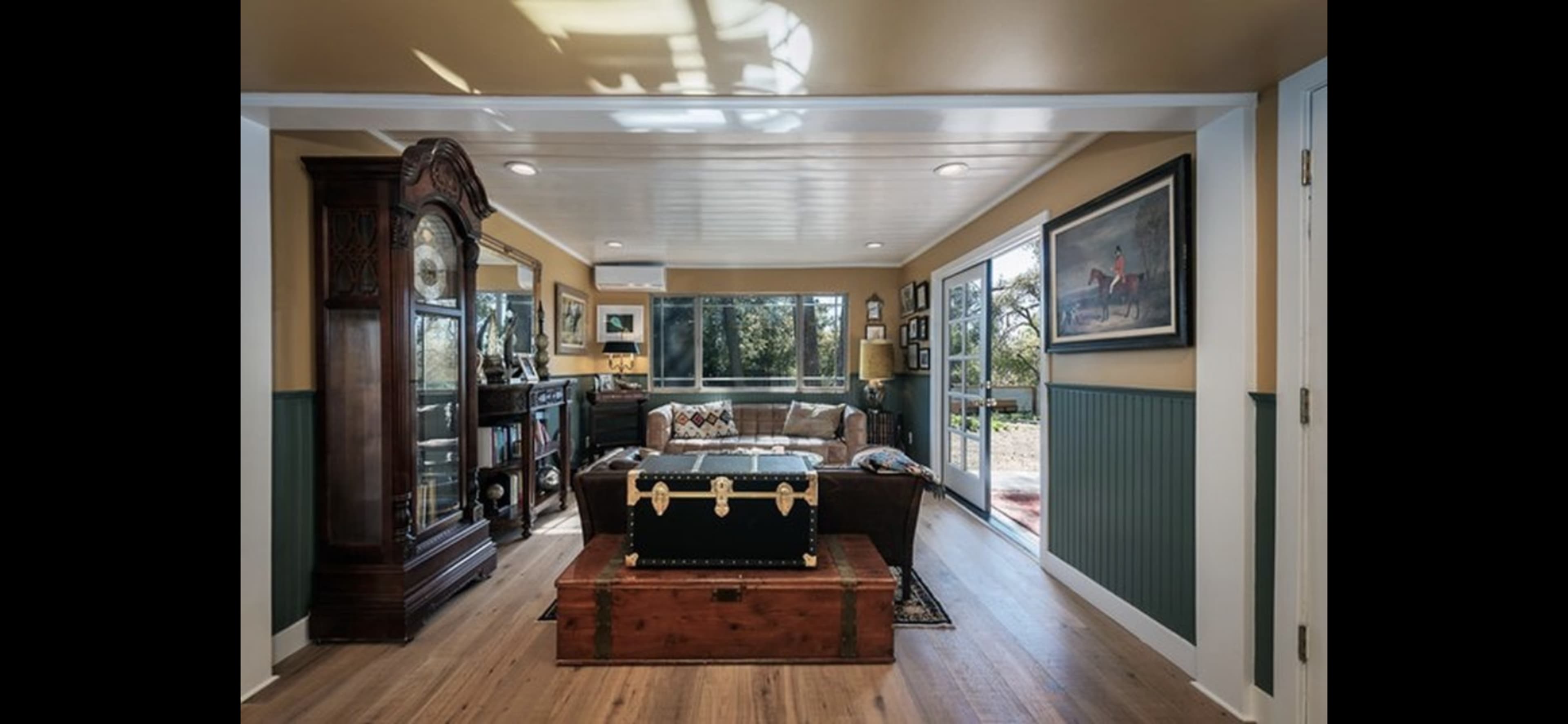 The image shows a well-lit living room featuring a wooden floor, a large window, a couch, and a decorative chest in the center.