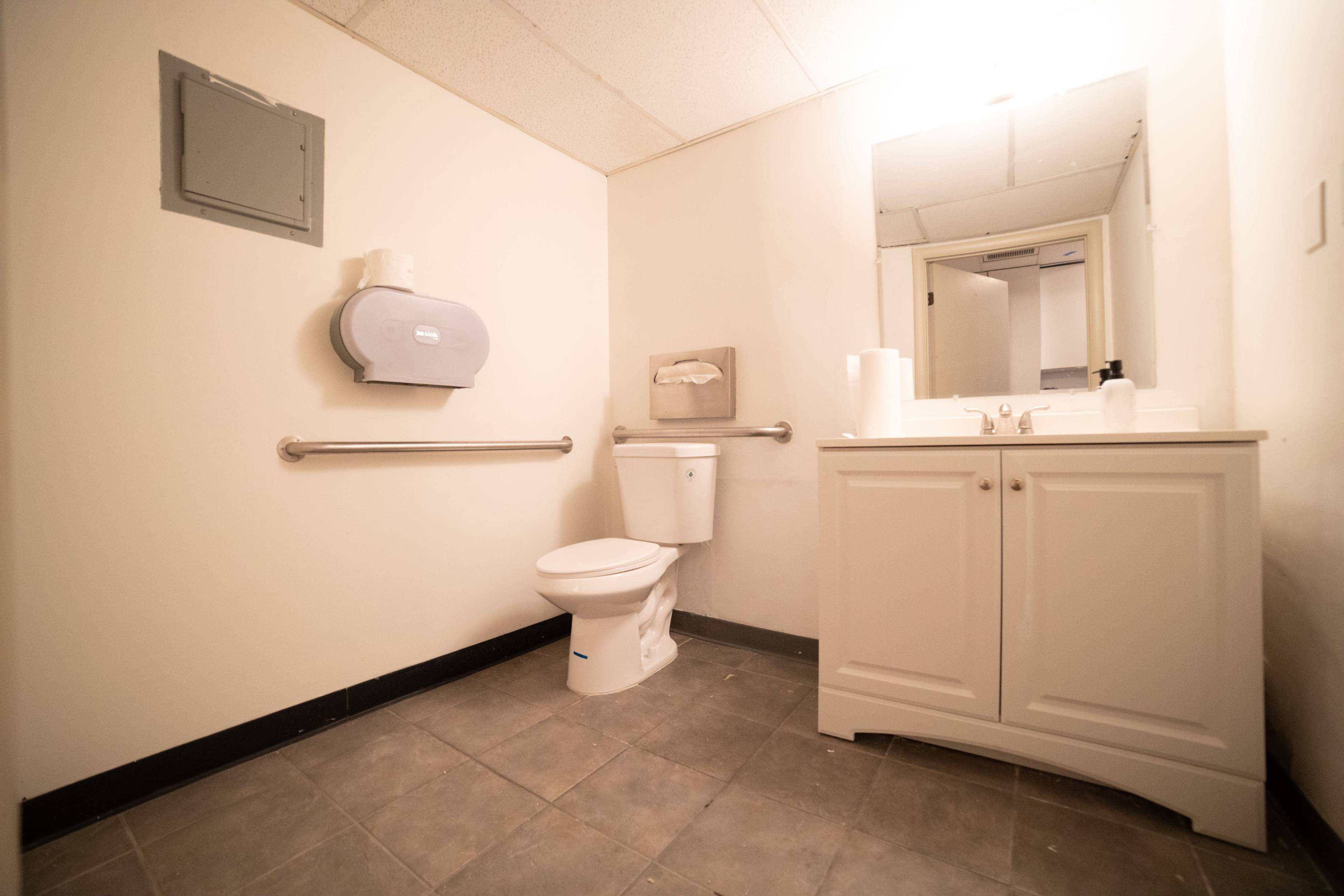 The image shows a restroom with a toilet, a sink with a cabinet, a paper towel dispenser, and a mirror on the wall.