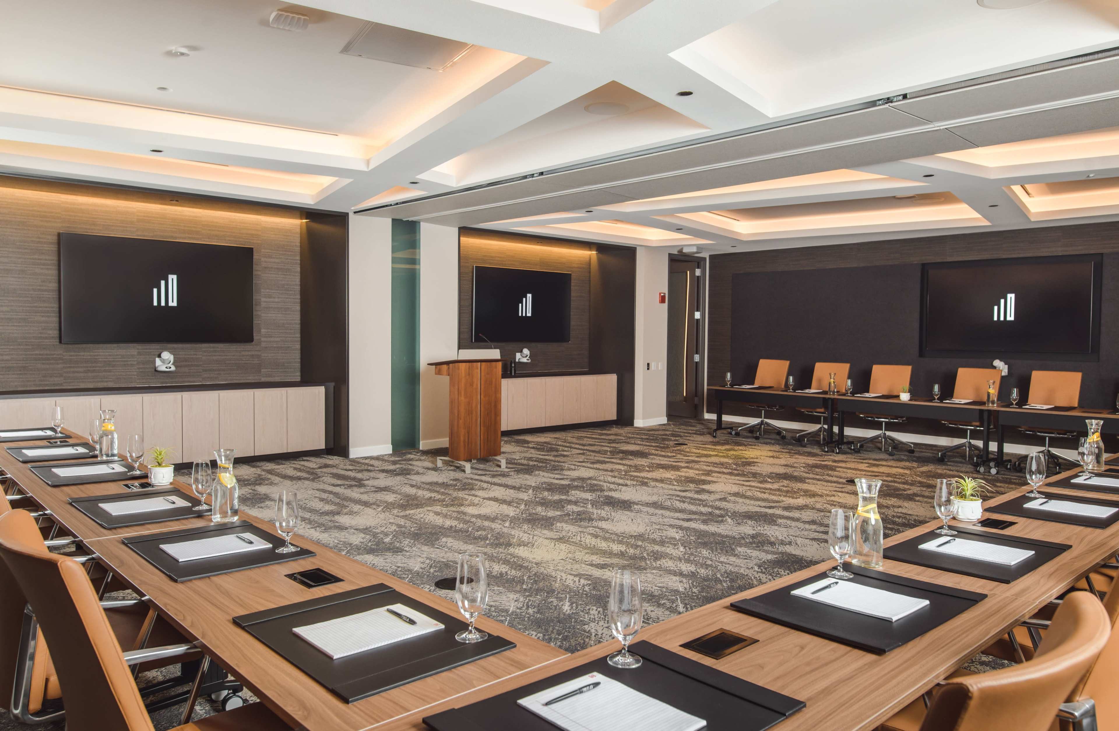 A modern conference room features a long table with place settings, two large screens on the walls, and a podium at the front.
