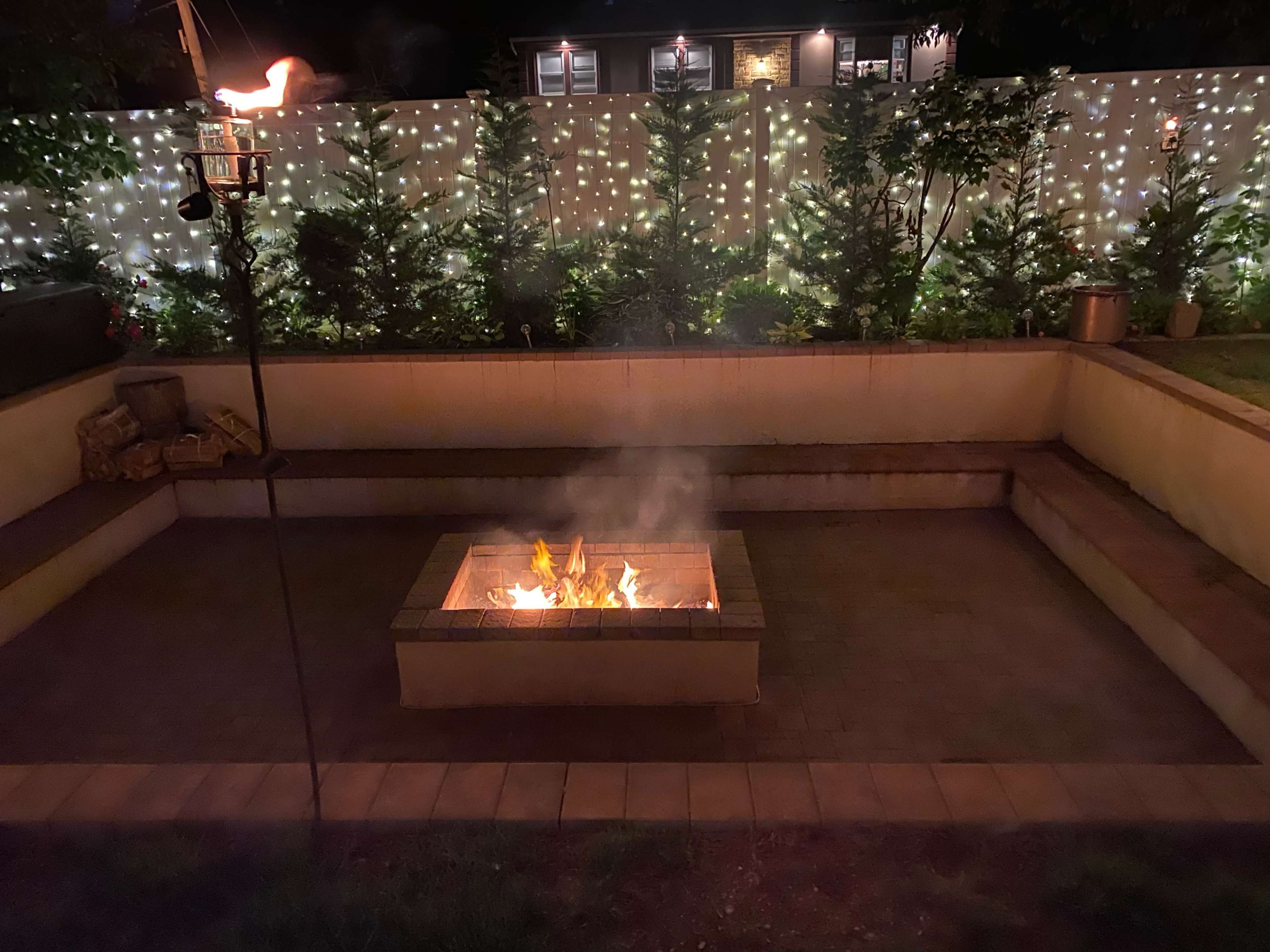 A fire burns in a stone fire pit surrounded by a paved seating area, with softly lit trees and a wall adorned with string lights in the background.