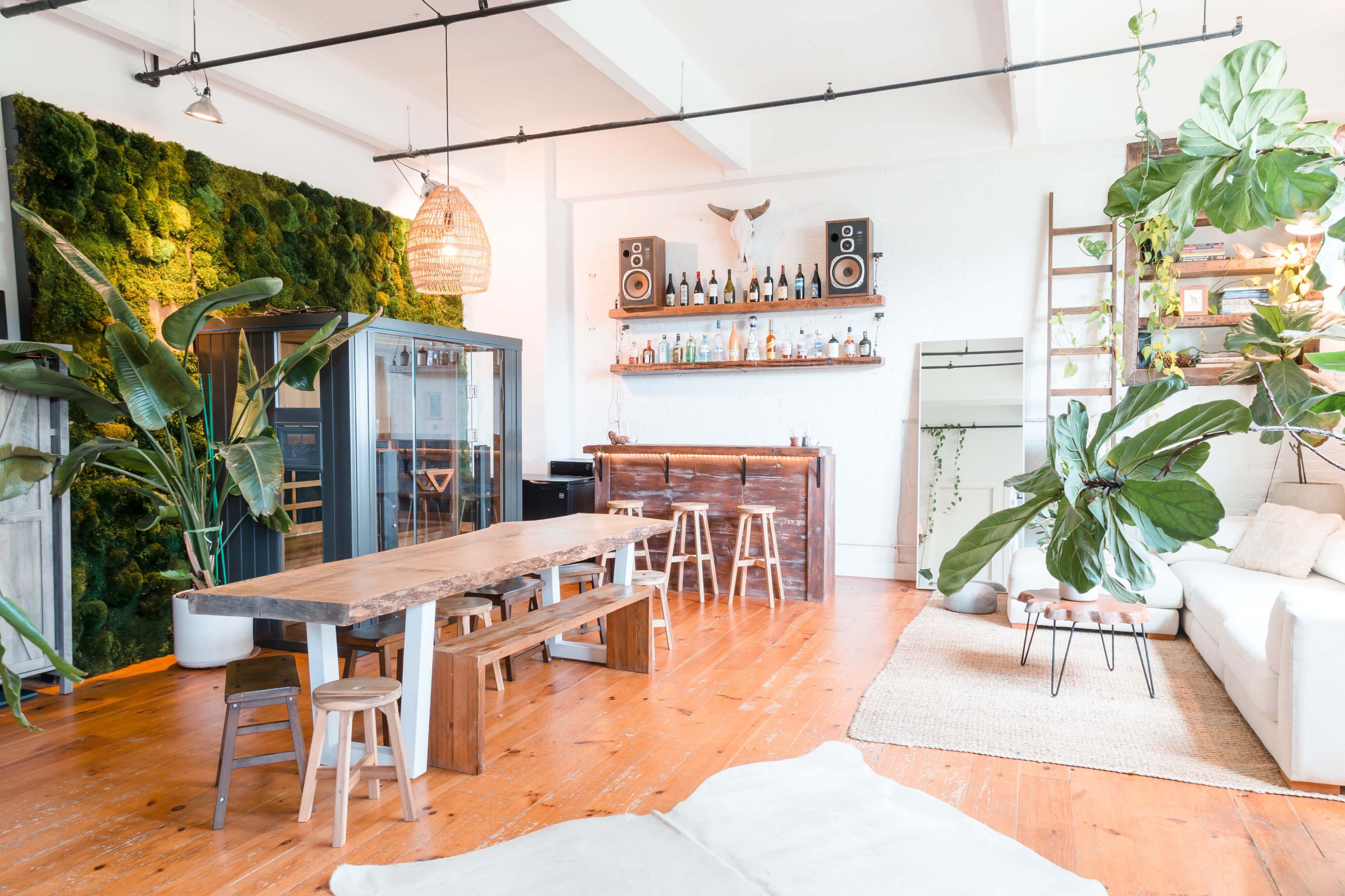 A spacious, modern interior featuring a wooden dining table, a green moss wall, and a bar area with bottles, surrounded by large plants and natural light.
