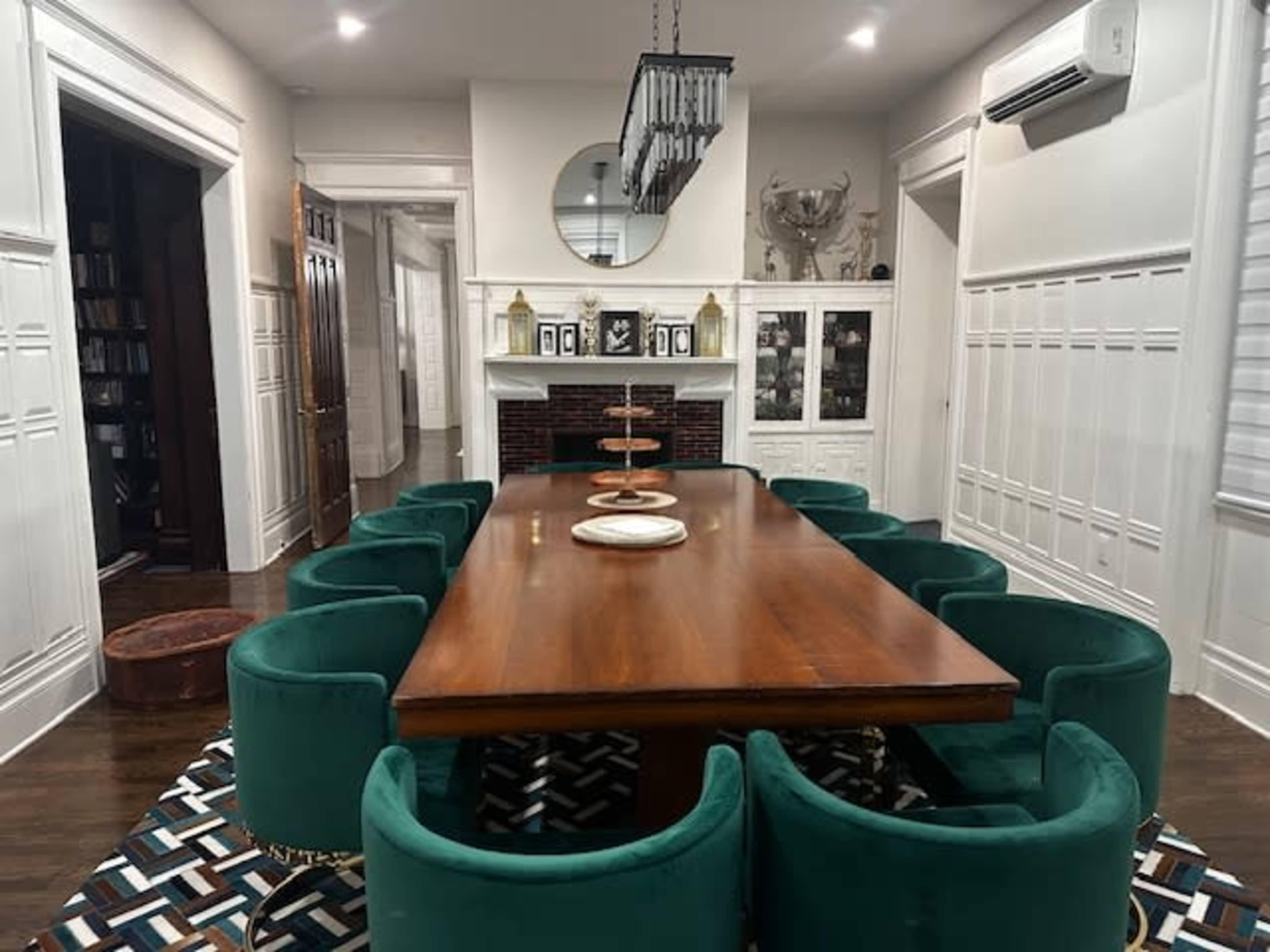 The image shows a dining room featuring a large wooden table surrounded by green upholstered chairs, with a patterned rug and decorative elements on the walls.
