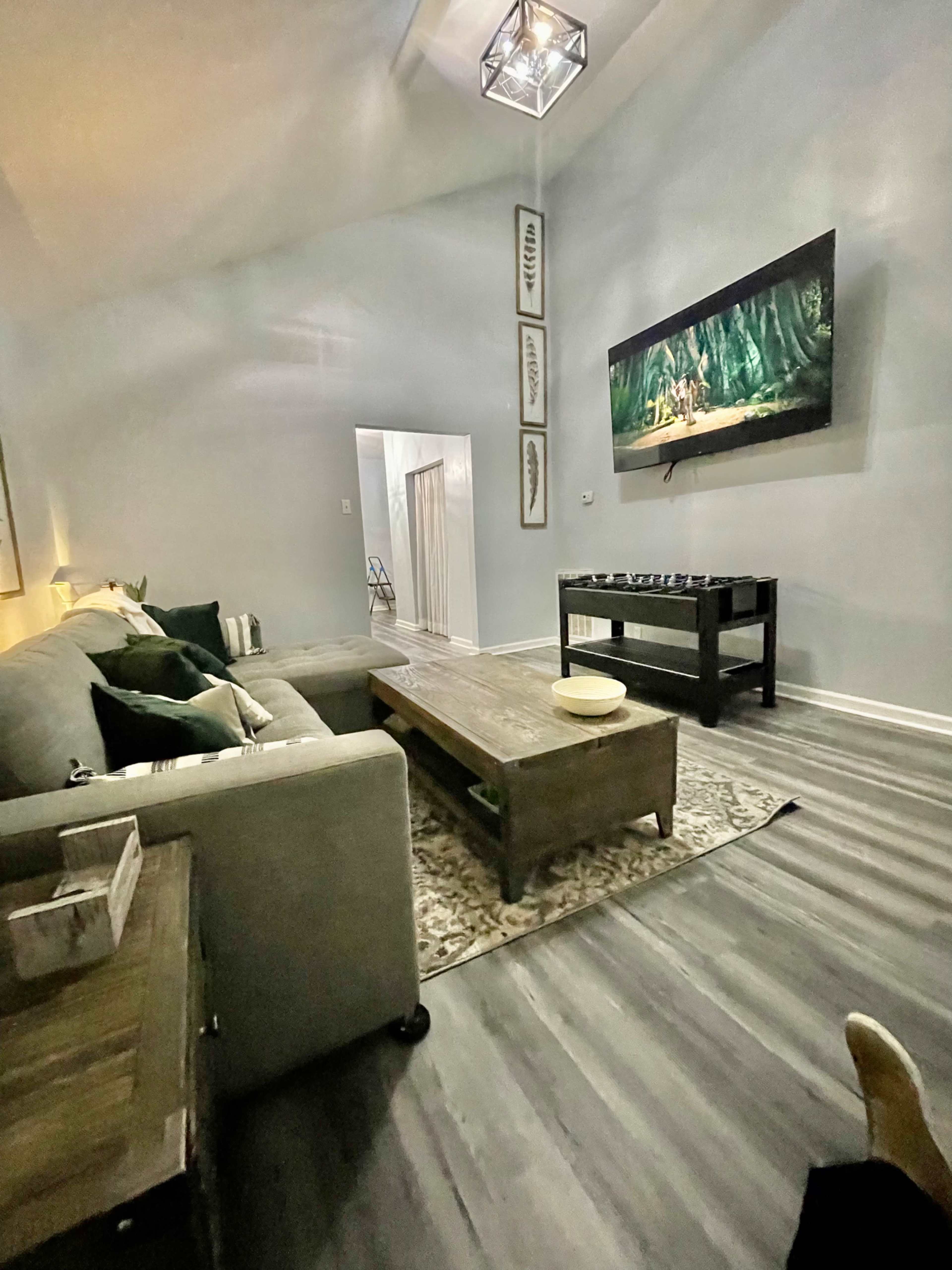 The living room features a gray sectional sofa, a wooden coffee table, and a television mounted on the wall, with light-colored walls and hardwood flooring.