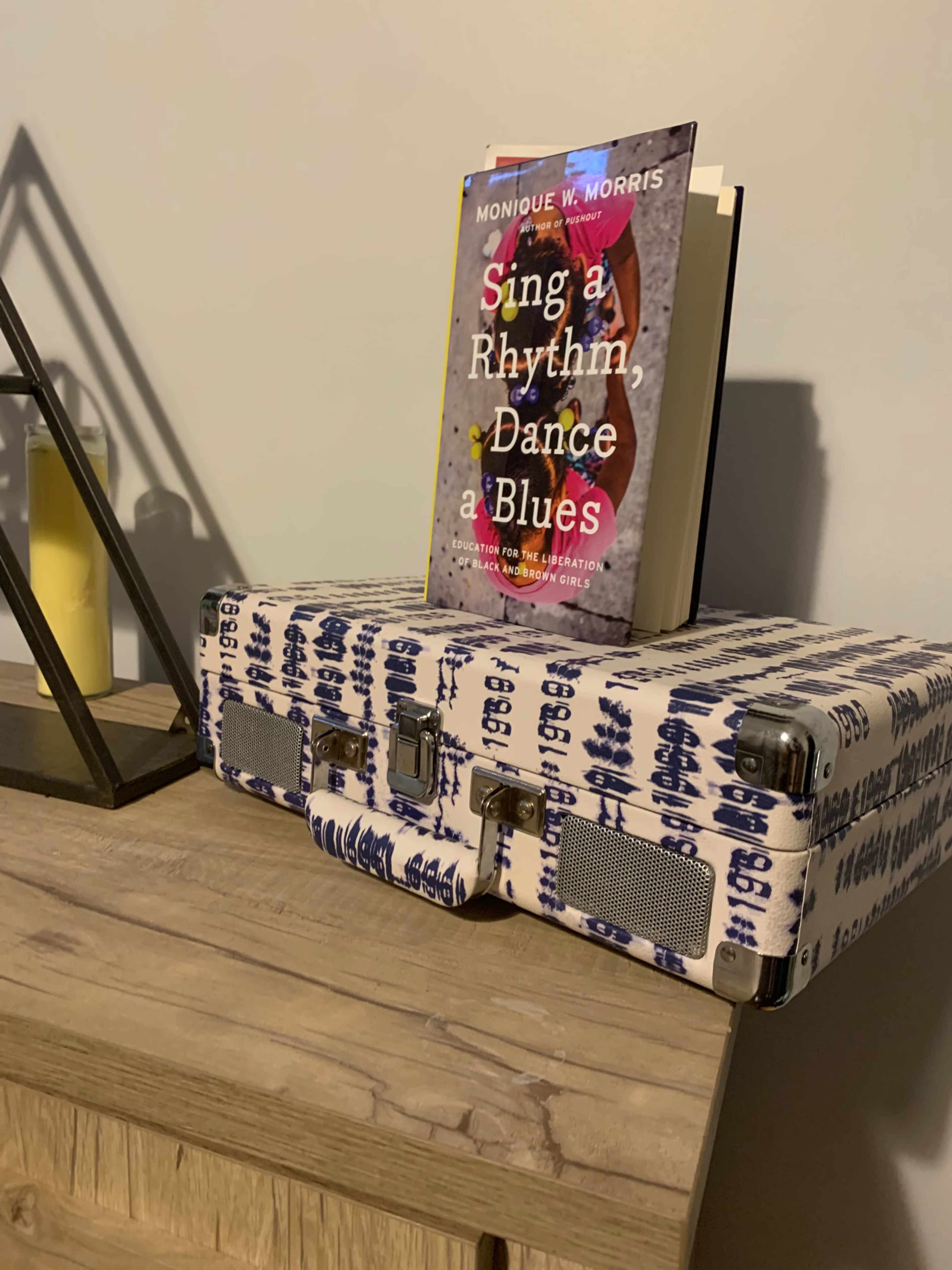 A book titled "Sing a Rhythm, Dance a Blues" sits atop a patterned suitcase on a wooden table with a lamp and candles in the background.