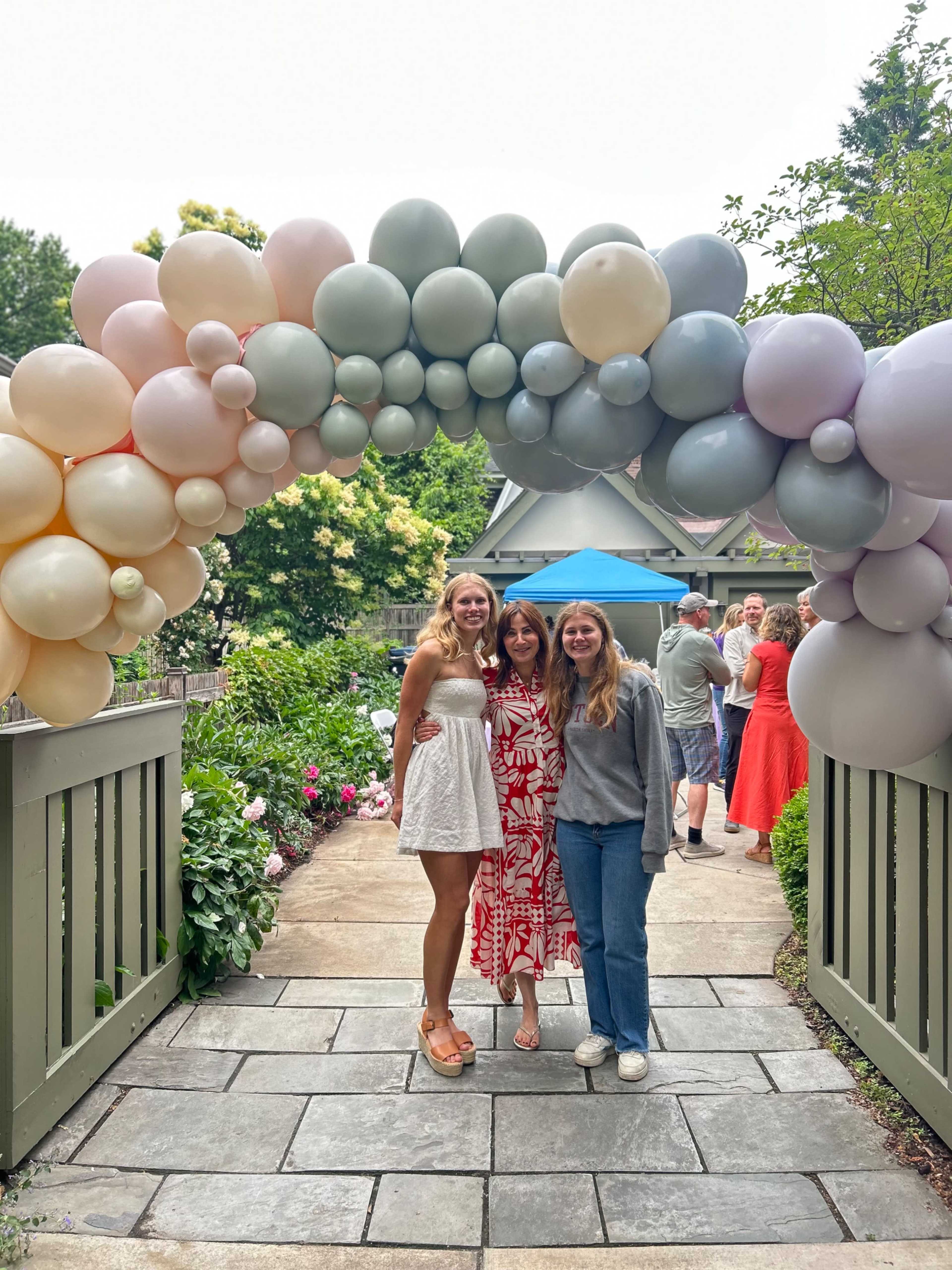 Three women pose under a balloon arch at a garden gathering, with guests mingling in the background.