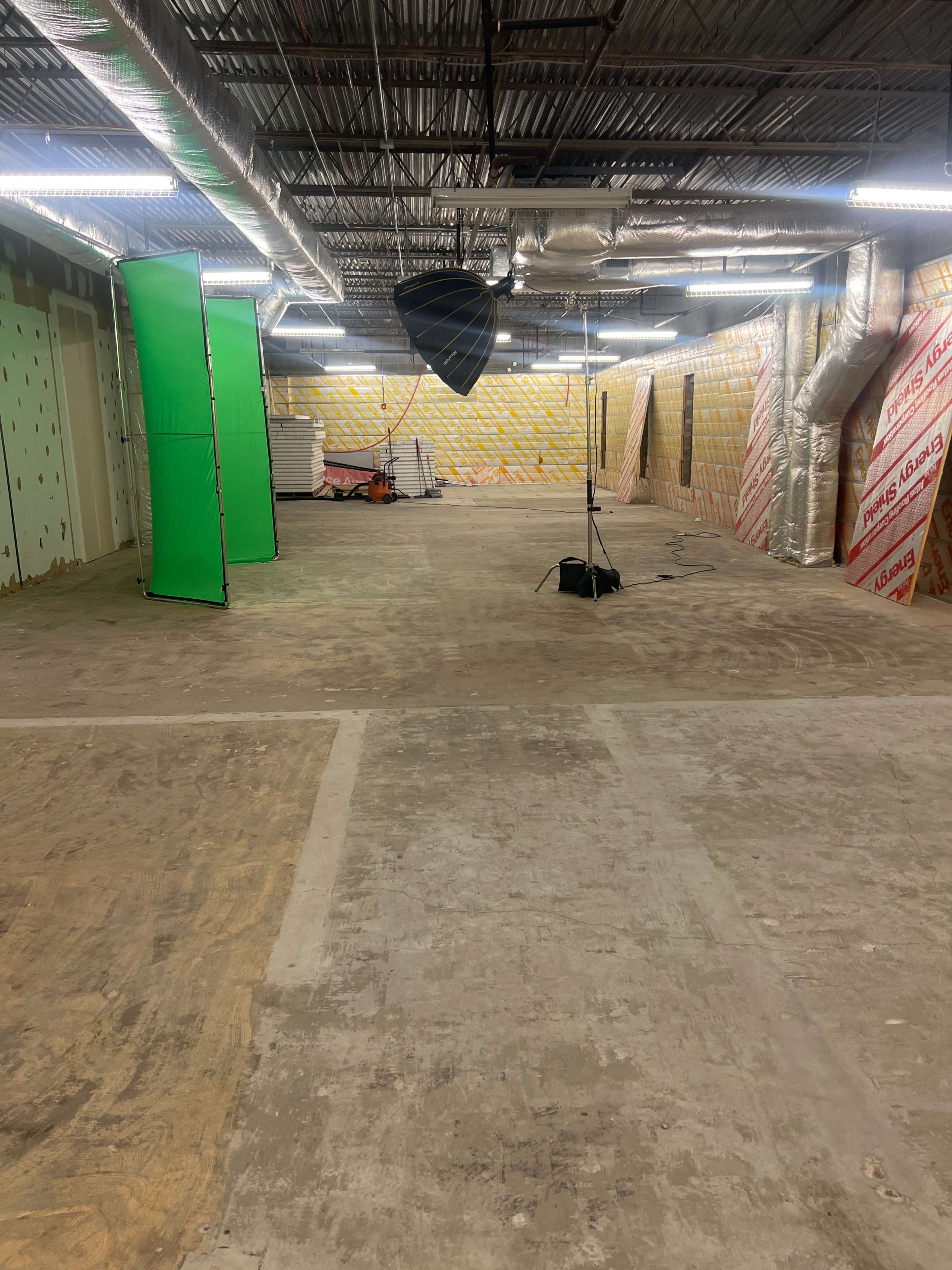 The image shows an empty, unfinished interior space with concrete flooring, green and black backdrop stands, and exposed ductwork on the ceiling.