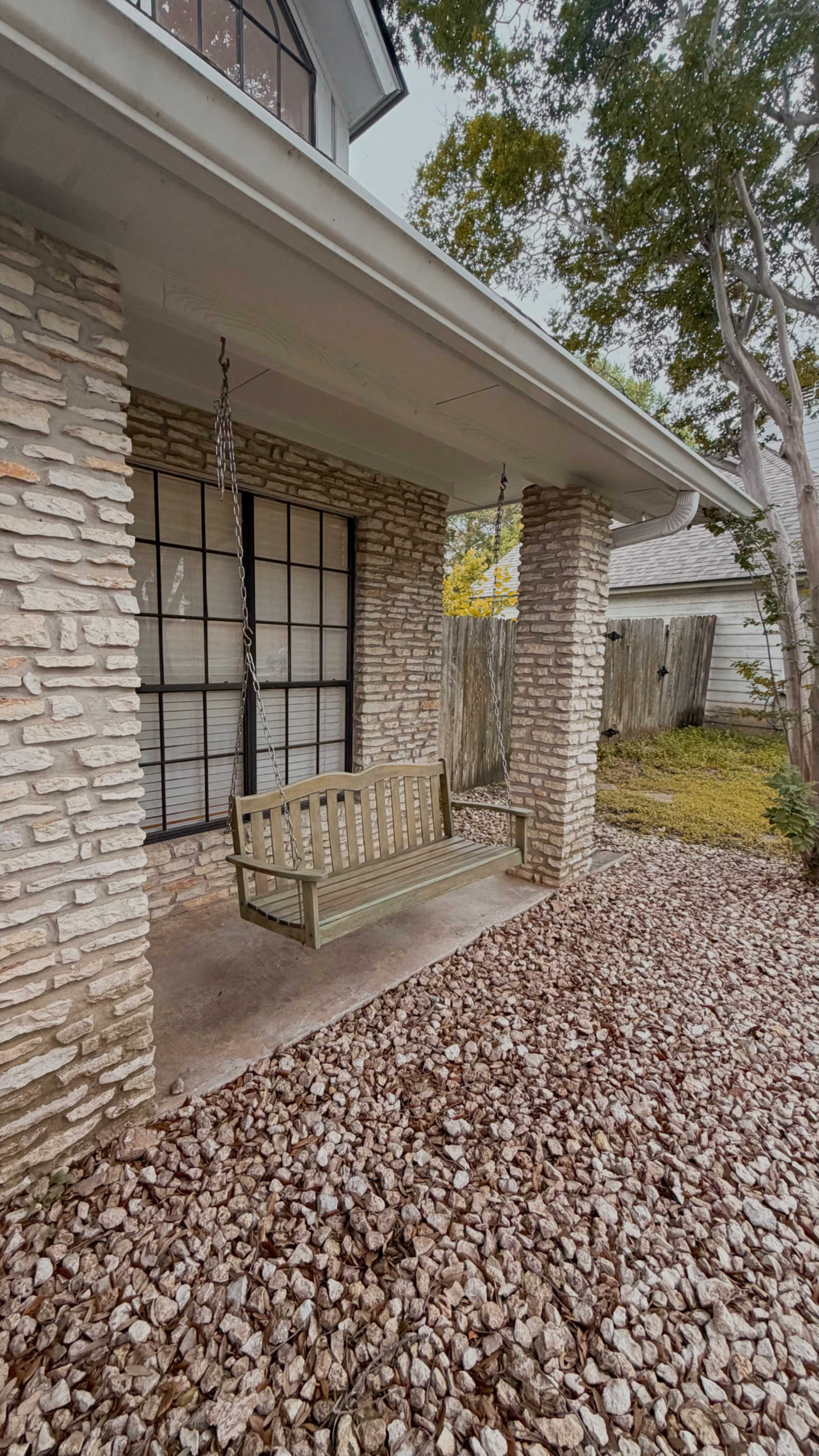 The image shows a covered porch with a wooden swing and stone pillars, surrounded by gravel and trees.