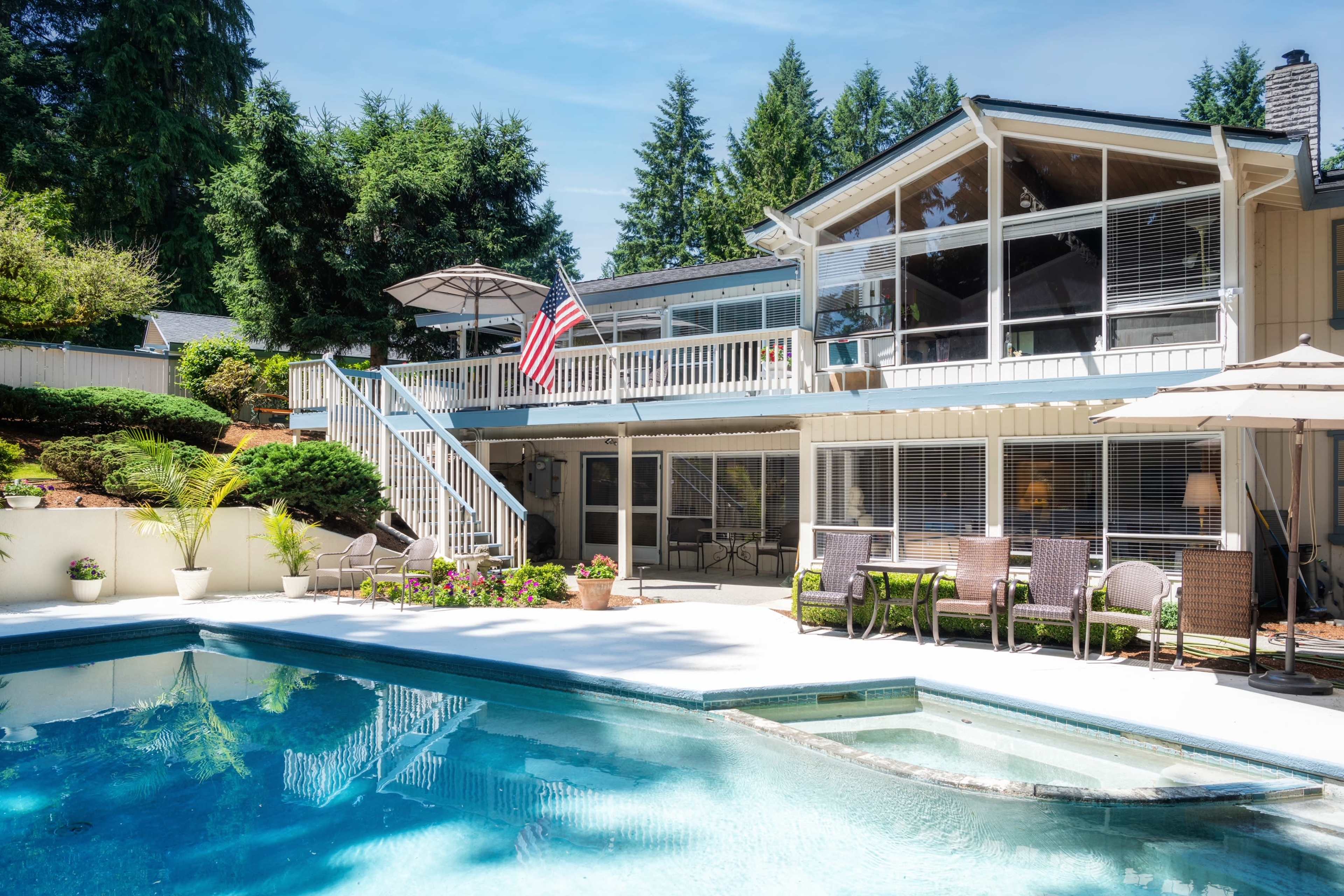 A large pool area is surrounded by a well-maintained patio and a two-story house with an American flag, nestled among trees.