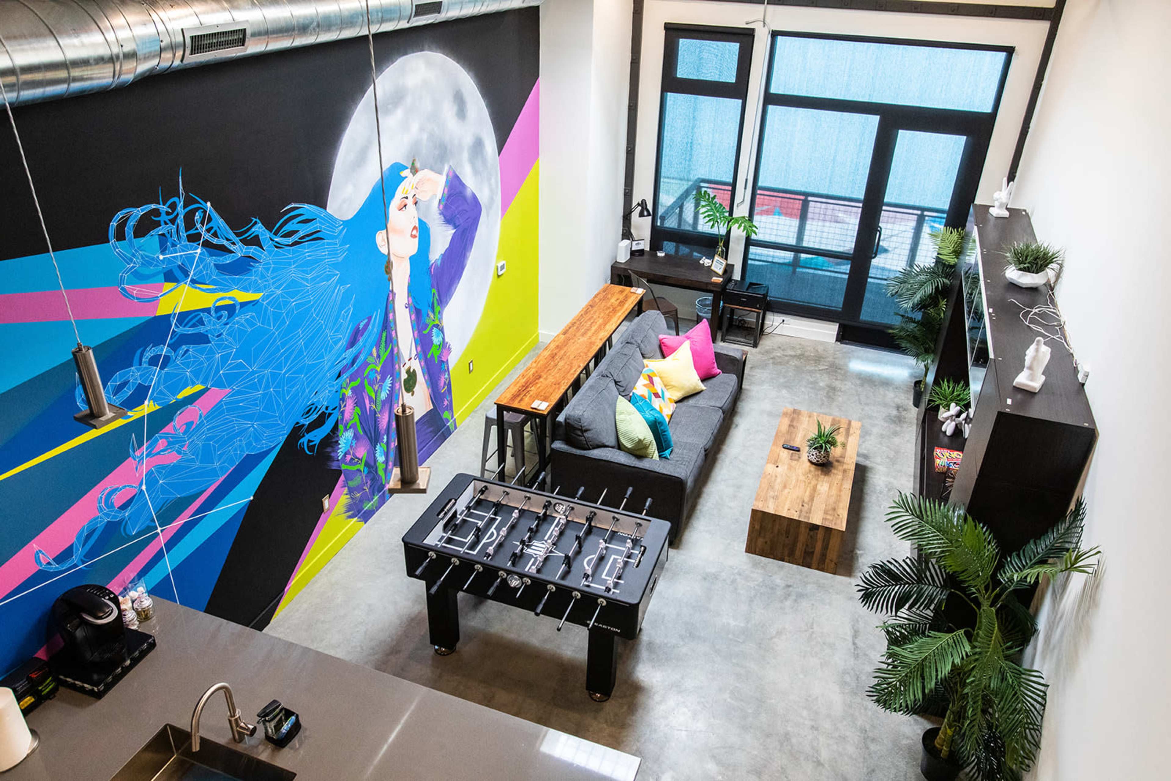 A modern living space with a large, colorful mural, a gray sofa with bright cushions, a wooden coffee table, a foosball table, and large windows letting in natural light.