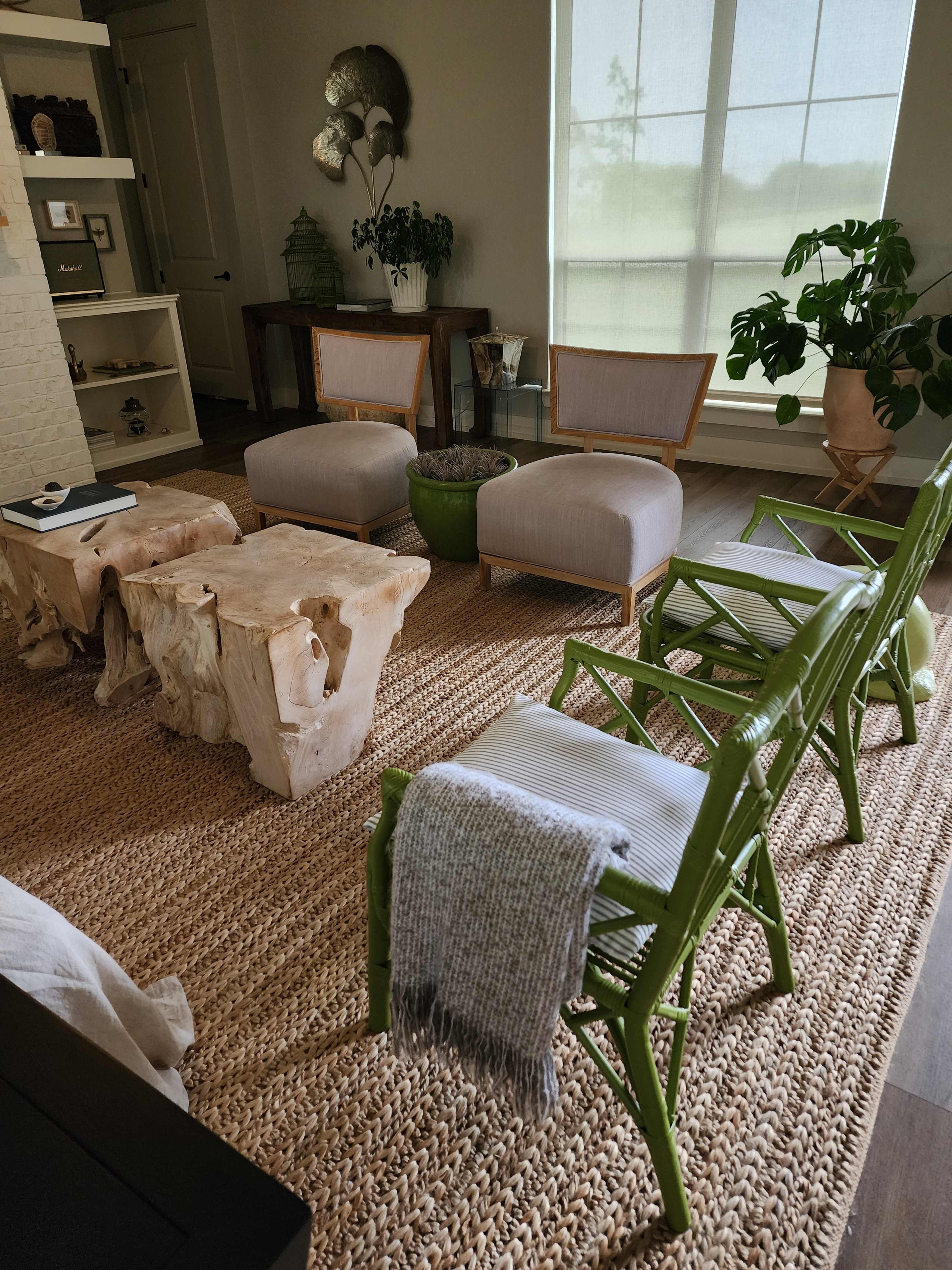 A modern living room features a seating area with two upholstered armchairs, a pair of stone side tables, and green wicker chairs on a textured rug.