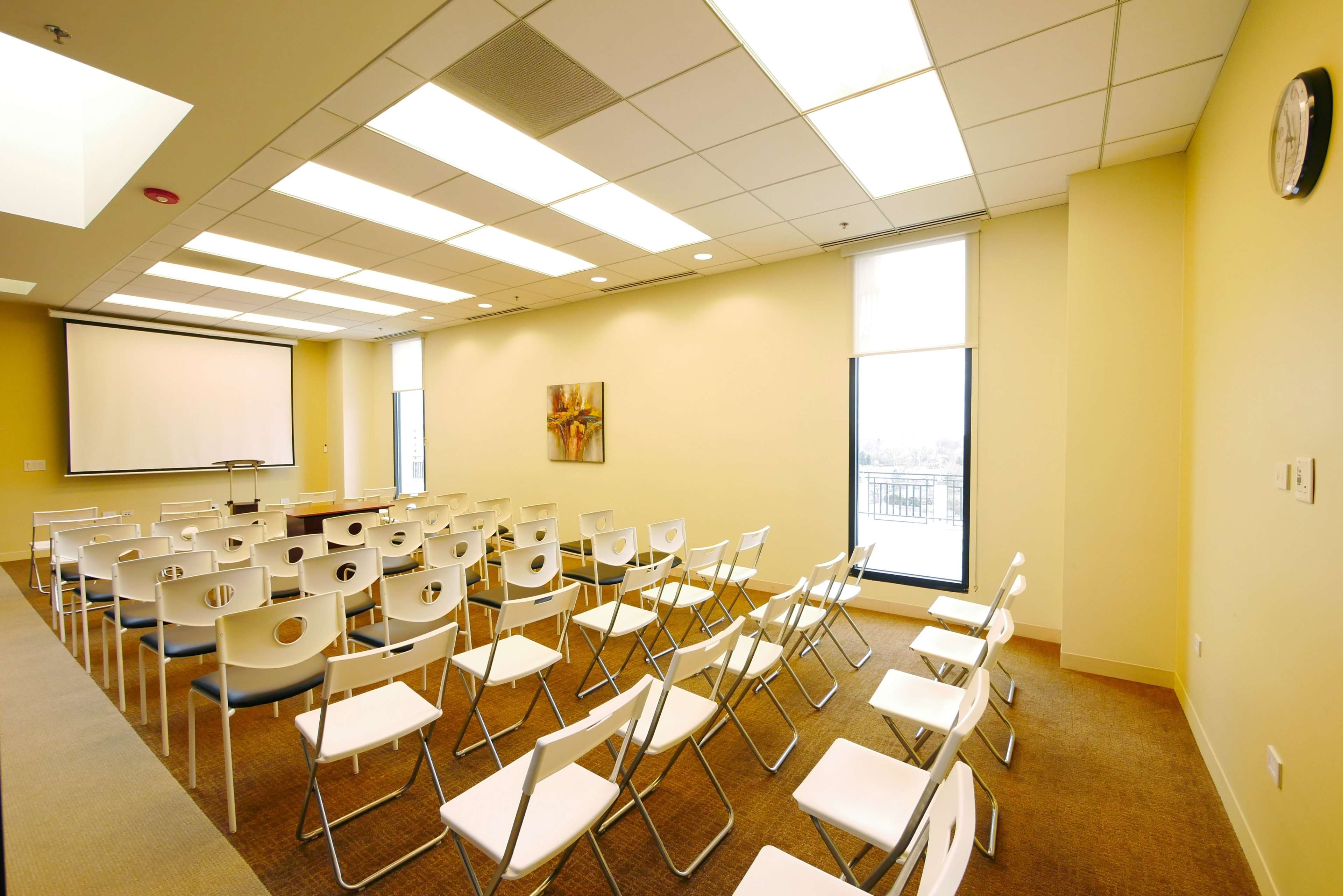 A conference room is set up with white chairs facing a blank projector screen and a large window.