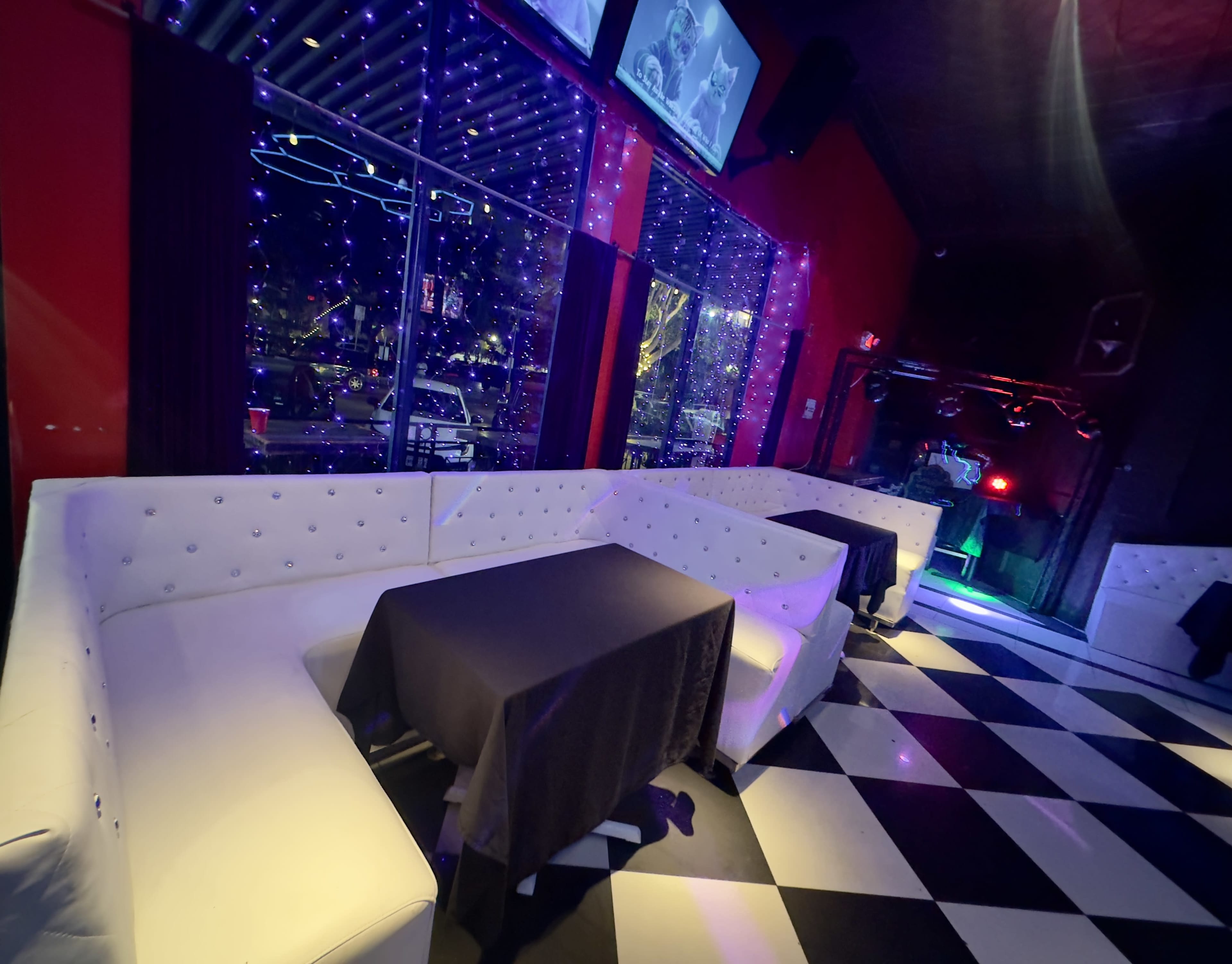 The image shows a lounge area with white seating, black tables, and a checkered floor, illuminated by colorful lights.