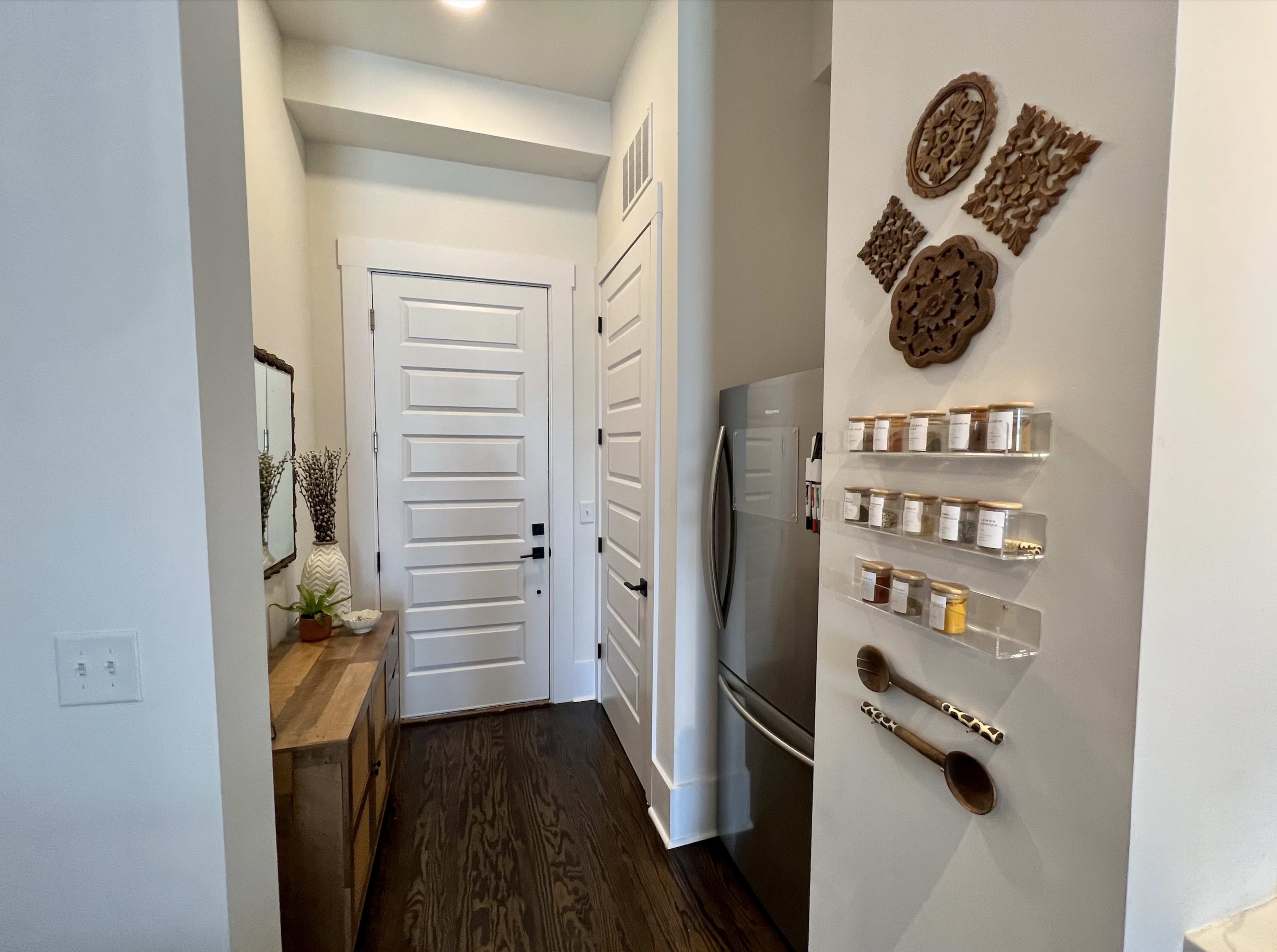 A well-lit entryway features a wooden console table, a mirror, and a wall-mounted shelf displaying jars and wooden utensils.