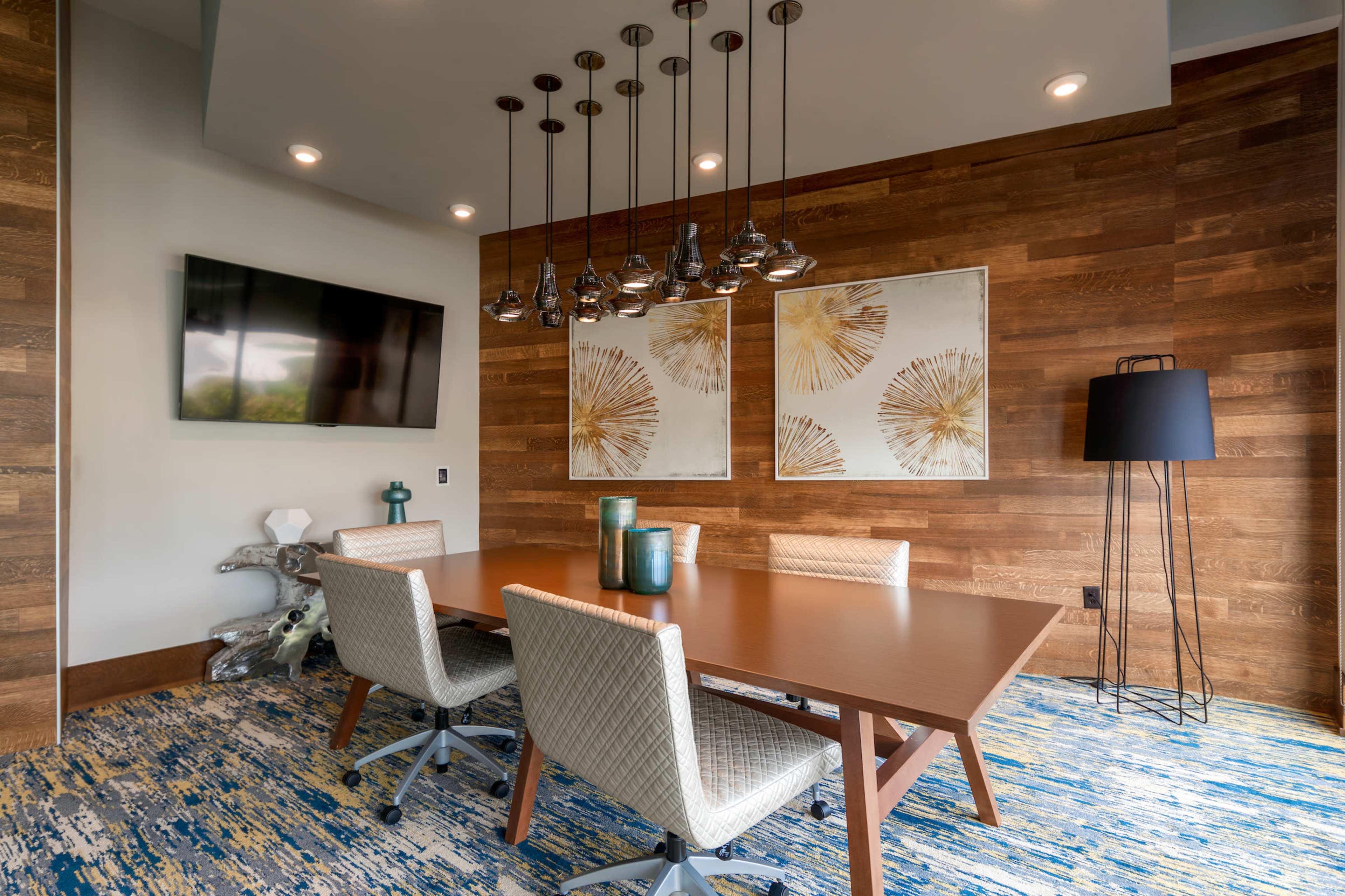A modern meeting room features a wooden table surrounded by upholstered chairs, with pendant lights hanging above and artwork on the walls.