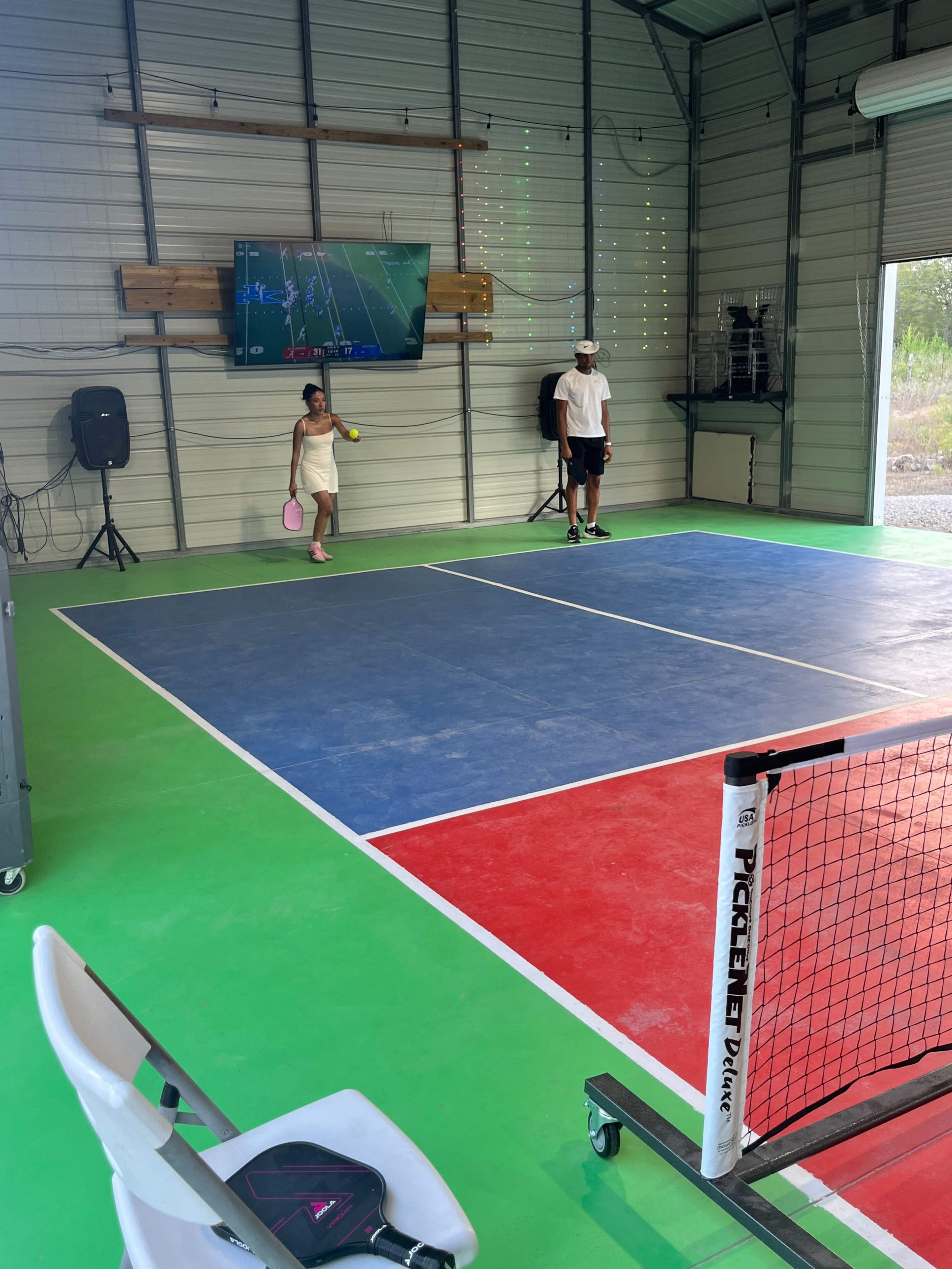 The image shows an indoor pickleball court with a monitor displaying a game, where a child is playing with a paddle while an adult observes.
