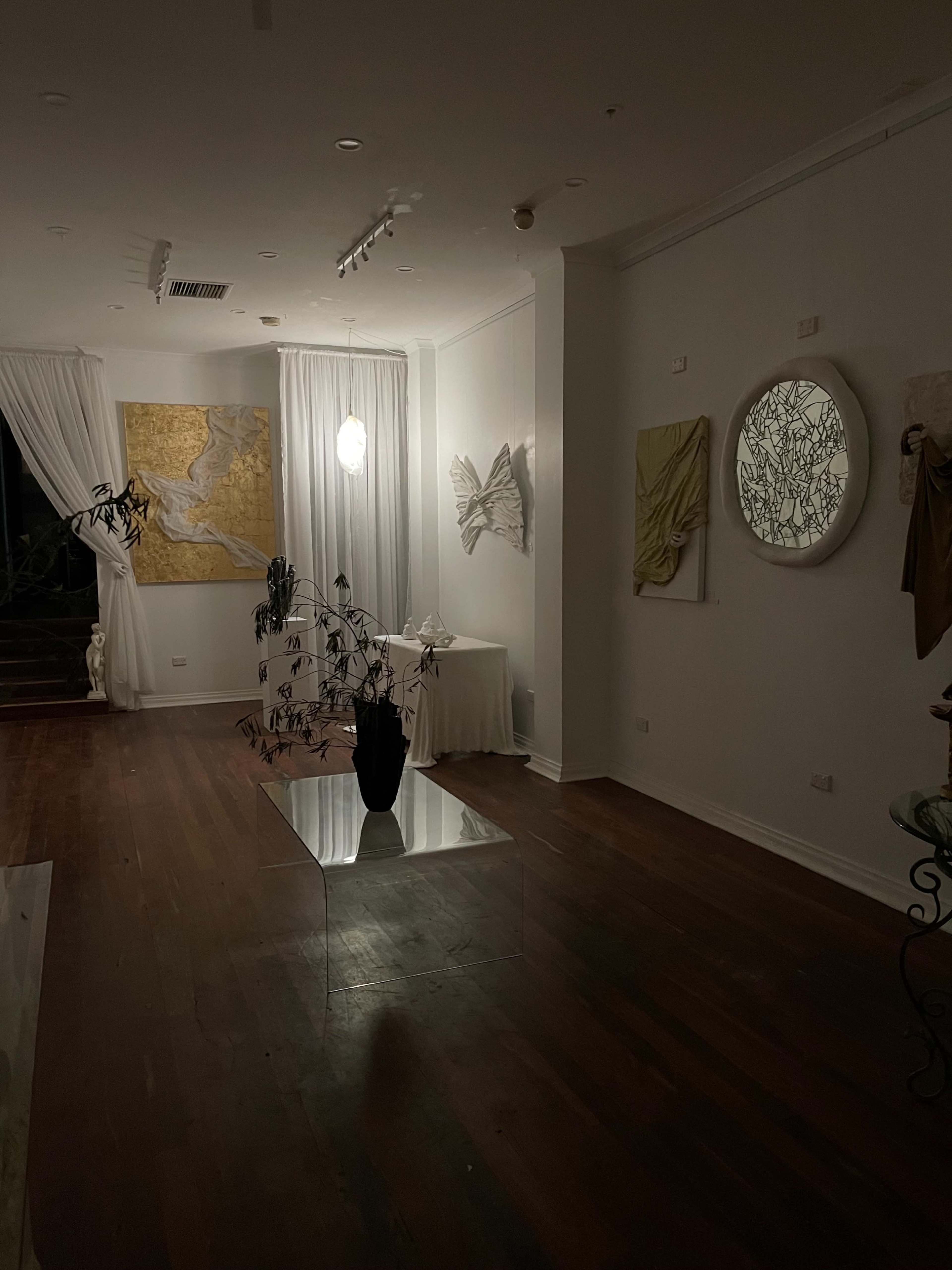 The image shows a dimly lit art gallery featuring white walls adorned with abstract artworks and a clear glass table with a plant arrangement.