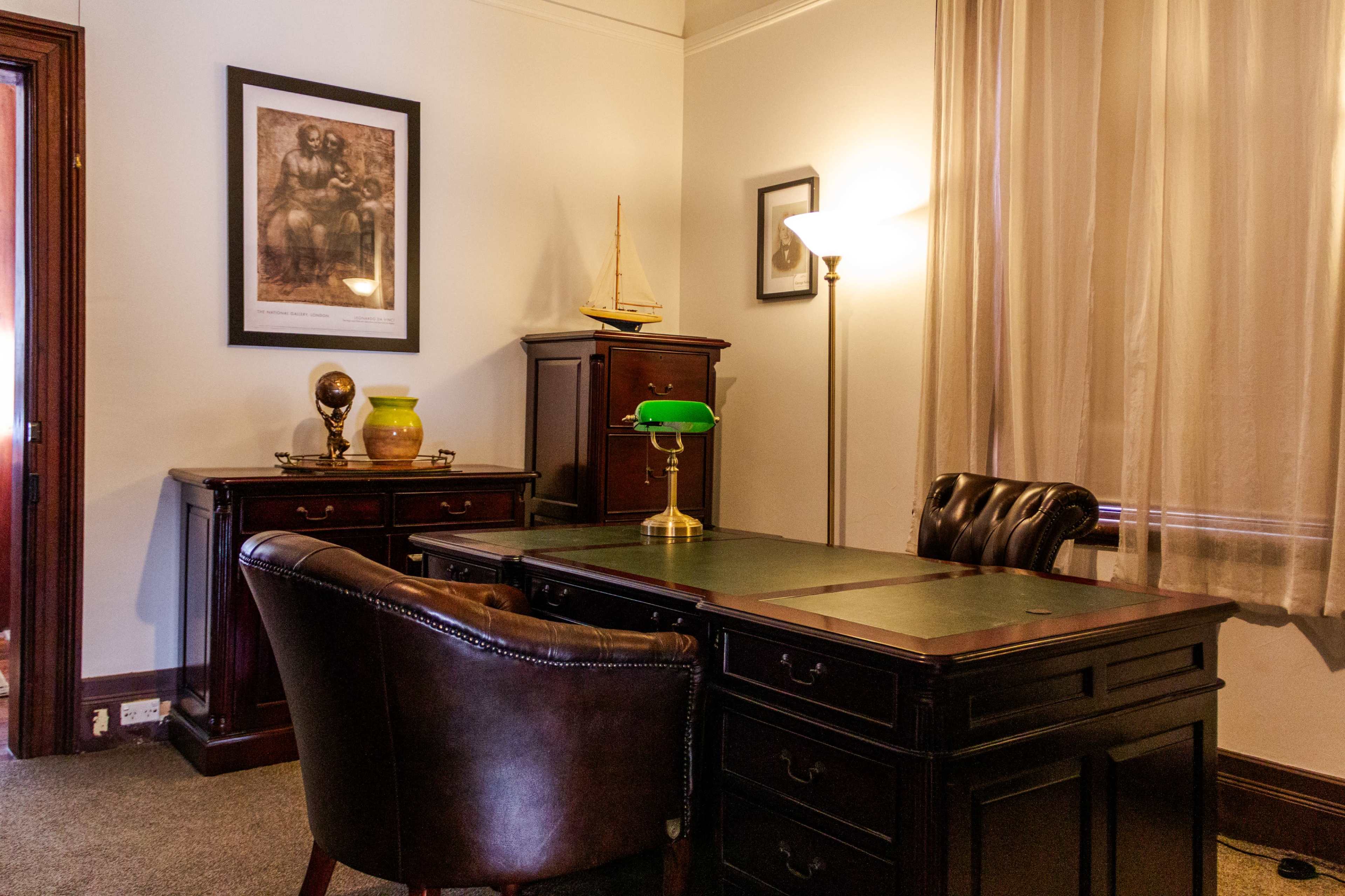 The image shows an elegantly furnished office with a wooden desk, leather chair, and vintage decor, including a lamp and a framed picture on the wall.