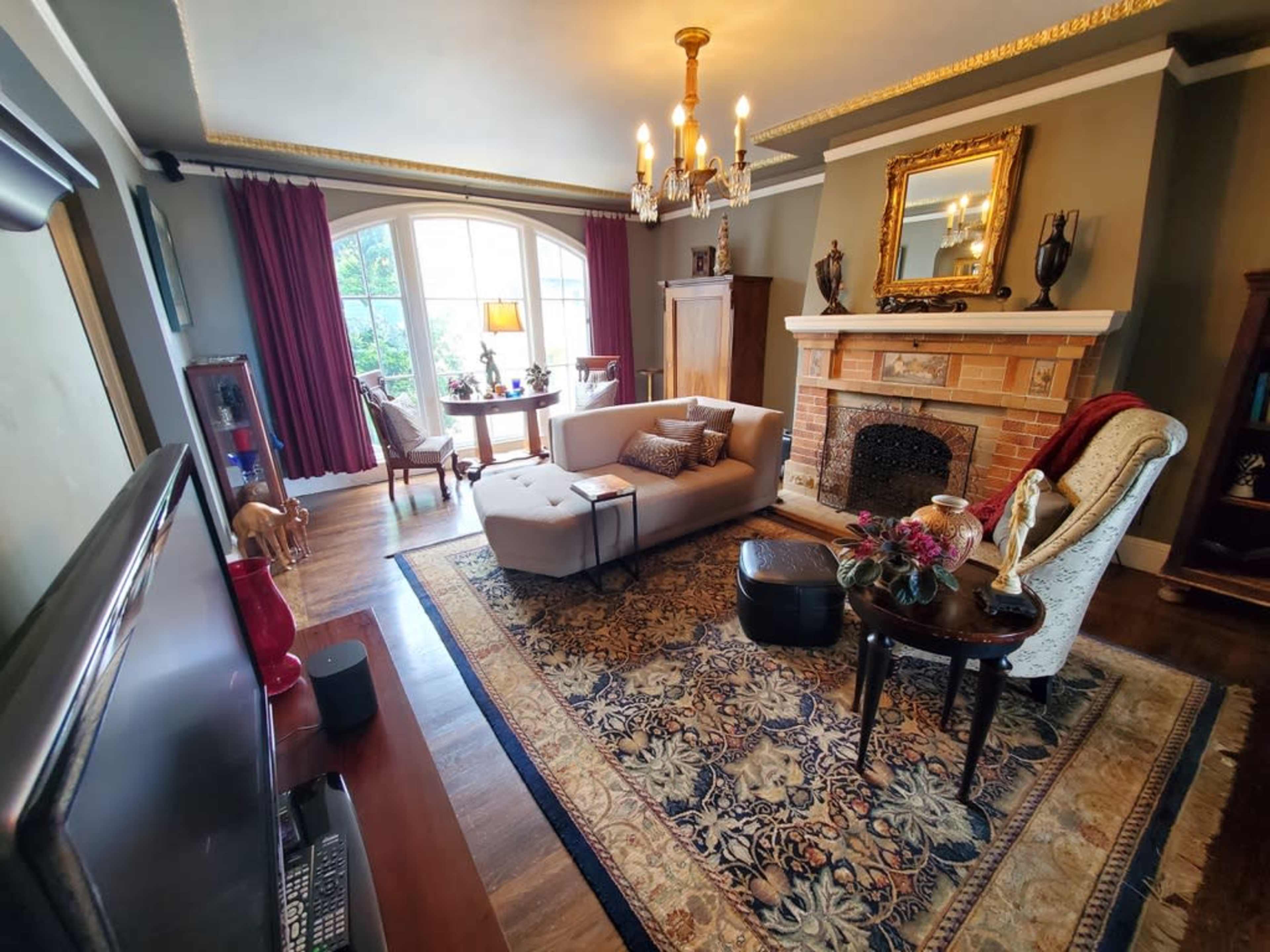 The image shows a cozy living room featuring a beige couch, vintage armchair, decorative fireplace, and a large window with red curtains, all set on a patterned area rug.
