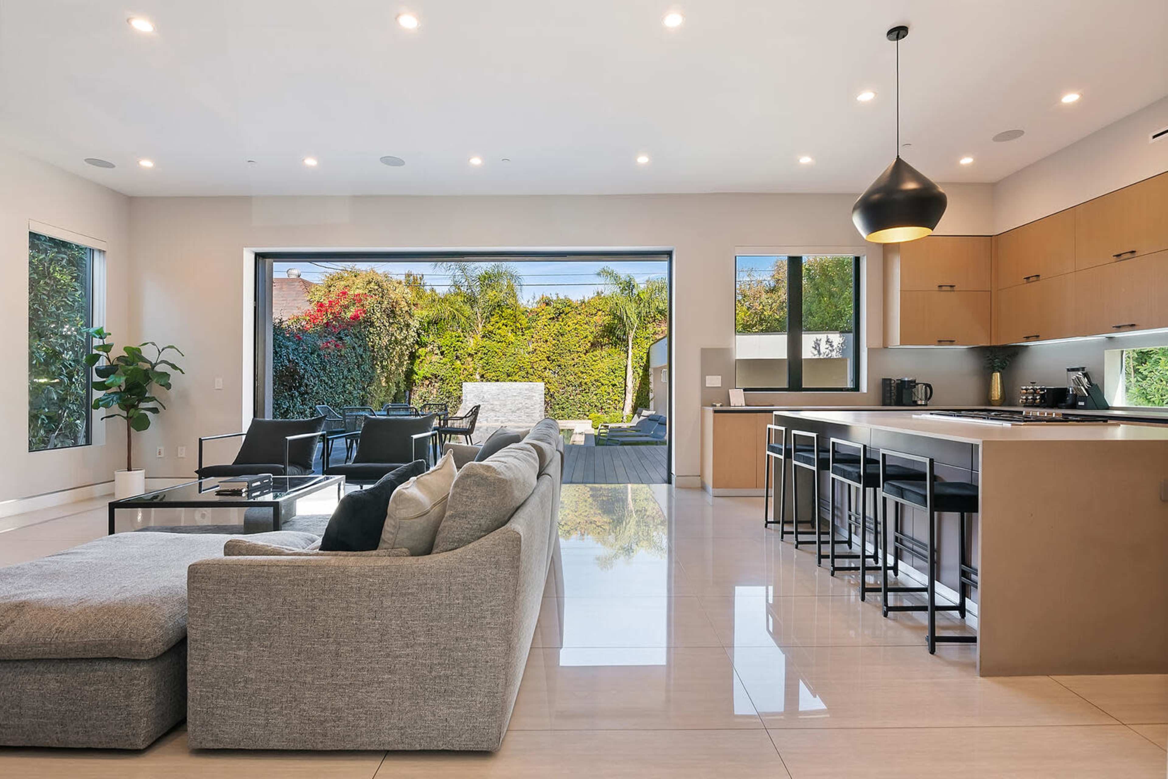 A modern open-concept living area featuring a gray sectional sofa, a kitchen with wooden cabinets, and large glass doors that lead to an outdoor patio.