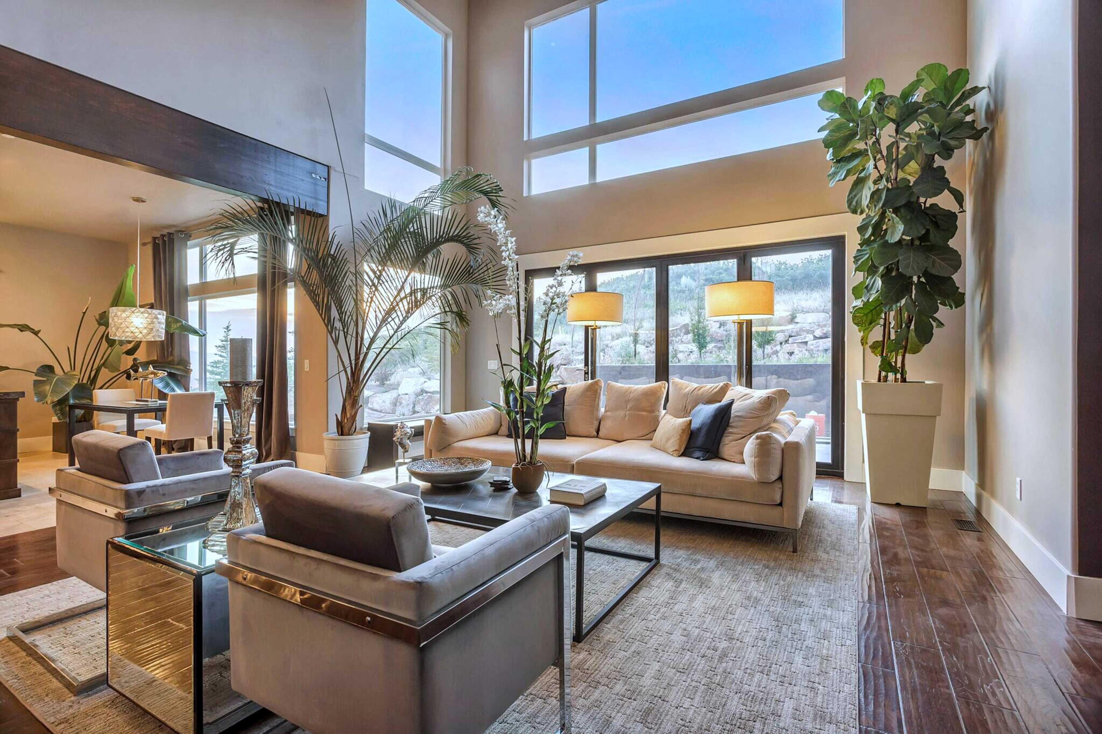 A modern living room with large windows, a light-colored sofa, two armchairs, and various indoor plants.