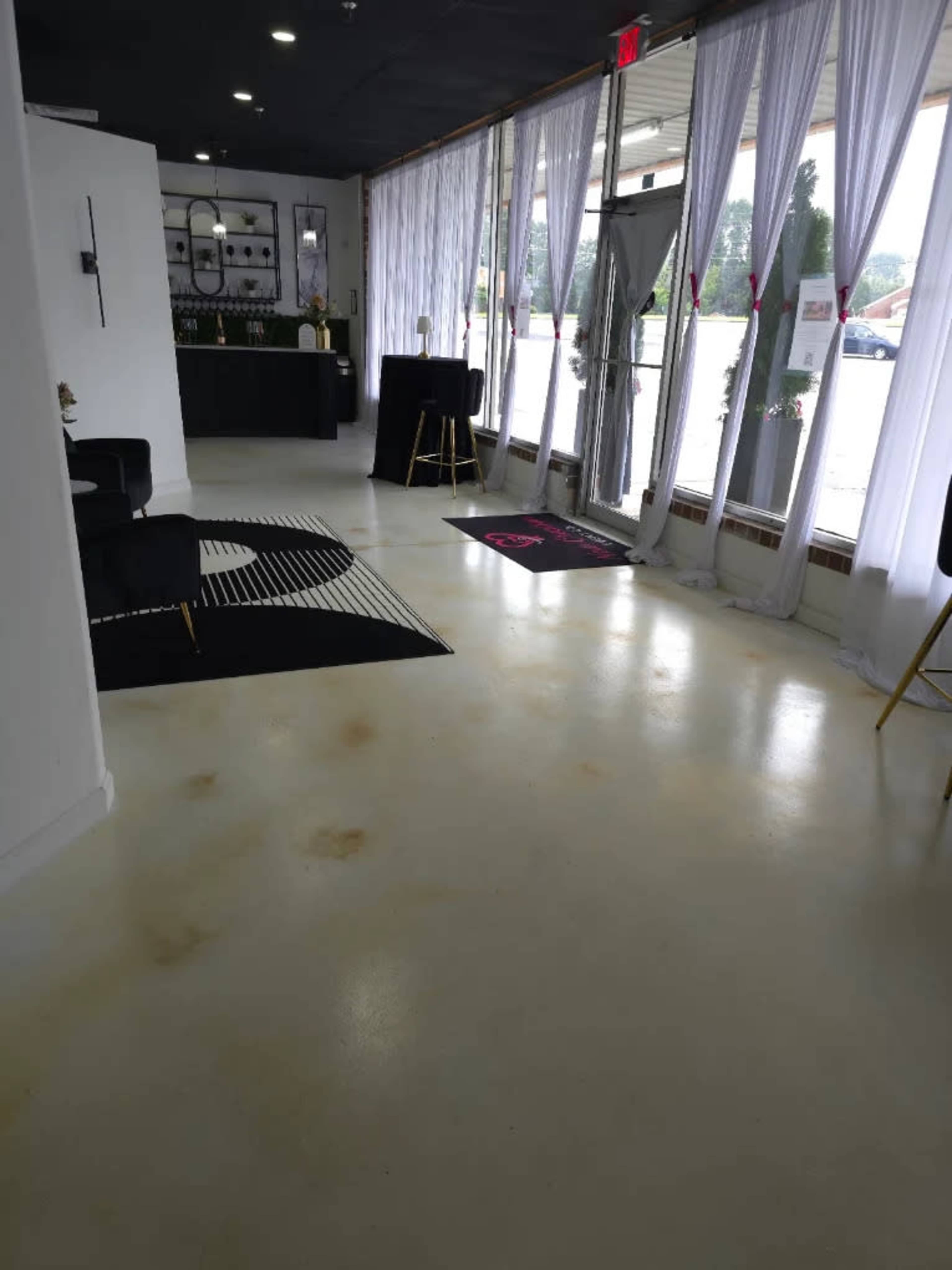 The image shows a spacious interior with large windows dressed in sheer curtains, a minimalist reception area with a dark counter, and a modern black and white rug on the floor.