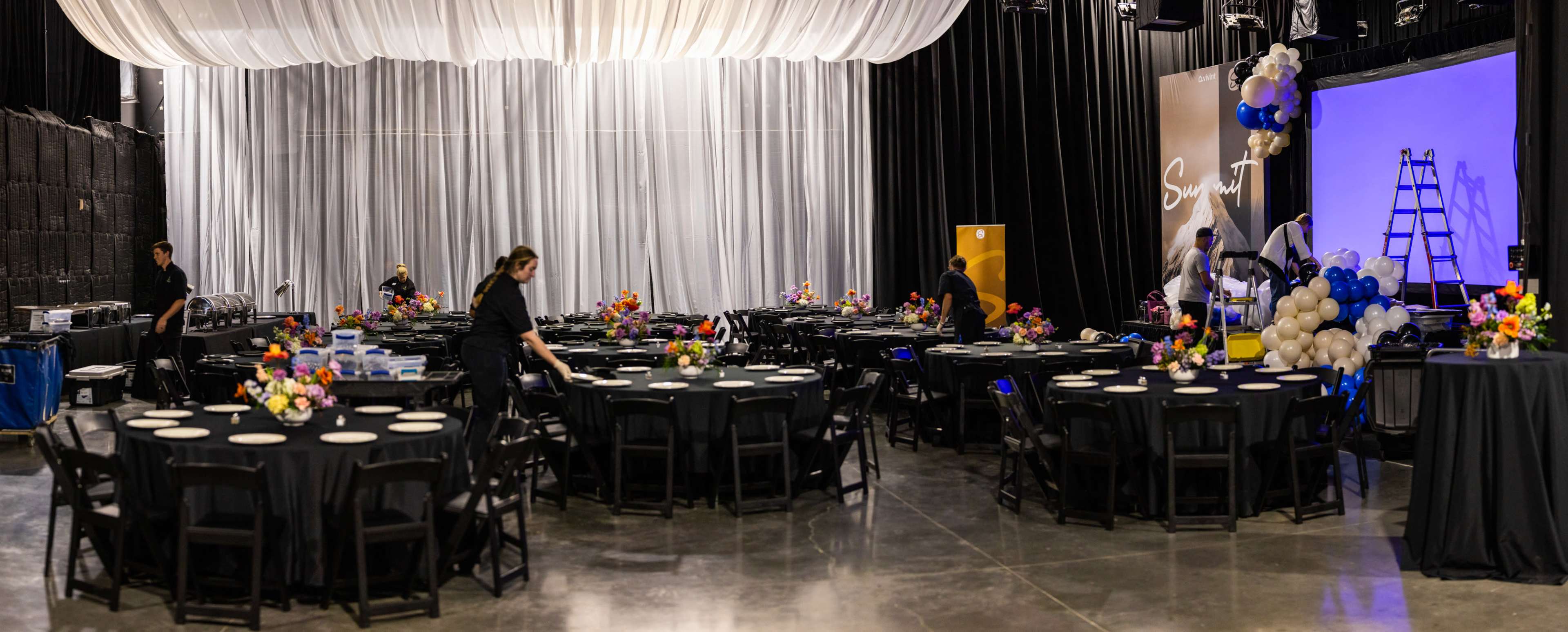 A spacious event venue is being prepared with black tables adorned with flower centerpieces, while staff members arrange the seating and decorations.